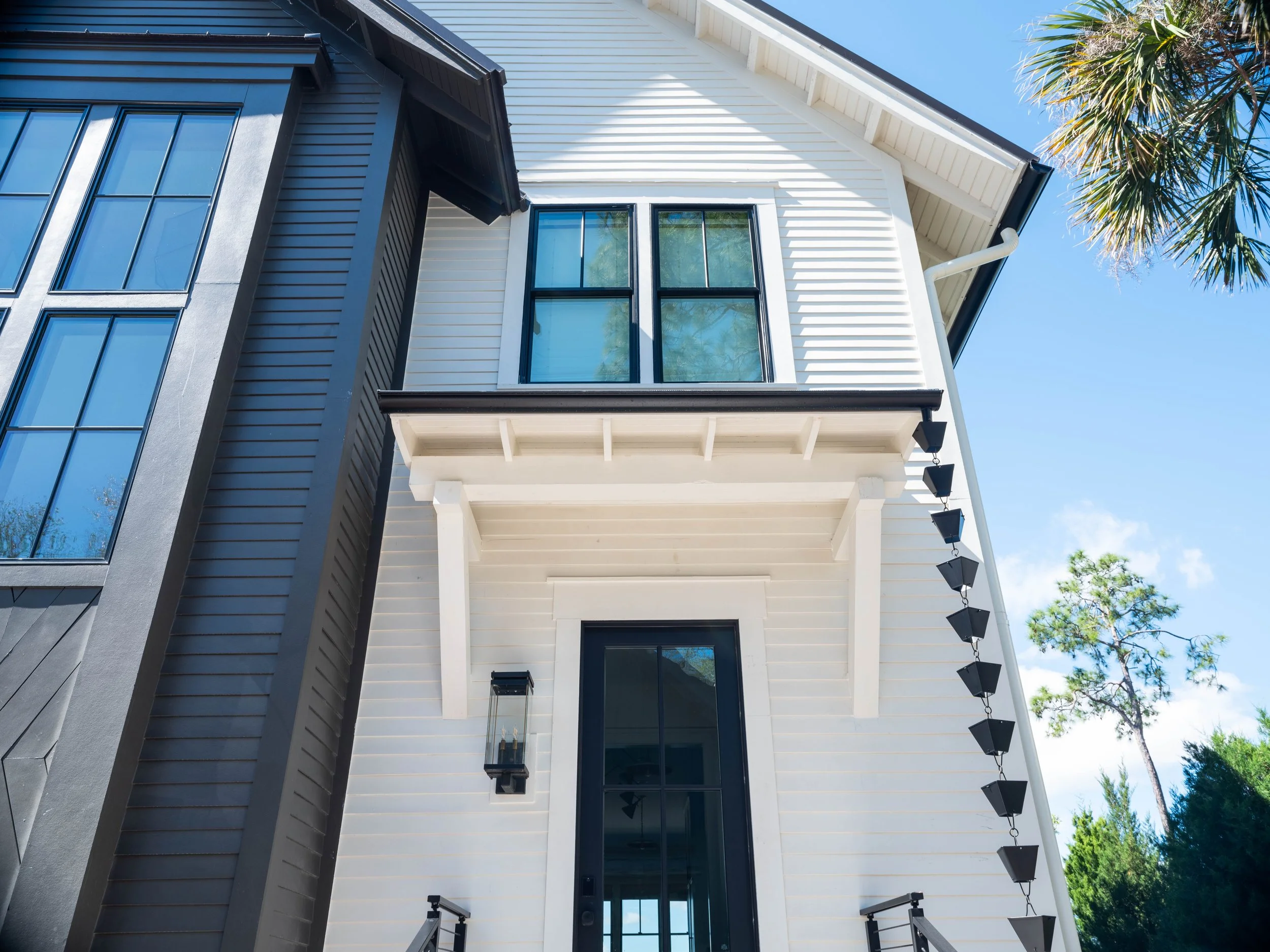 Crane Island Home. Exterior view of a modern house with white and gray siding, large windows, black accents, and a metal staircase with potted plants outside under a blue sky.