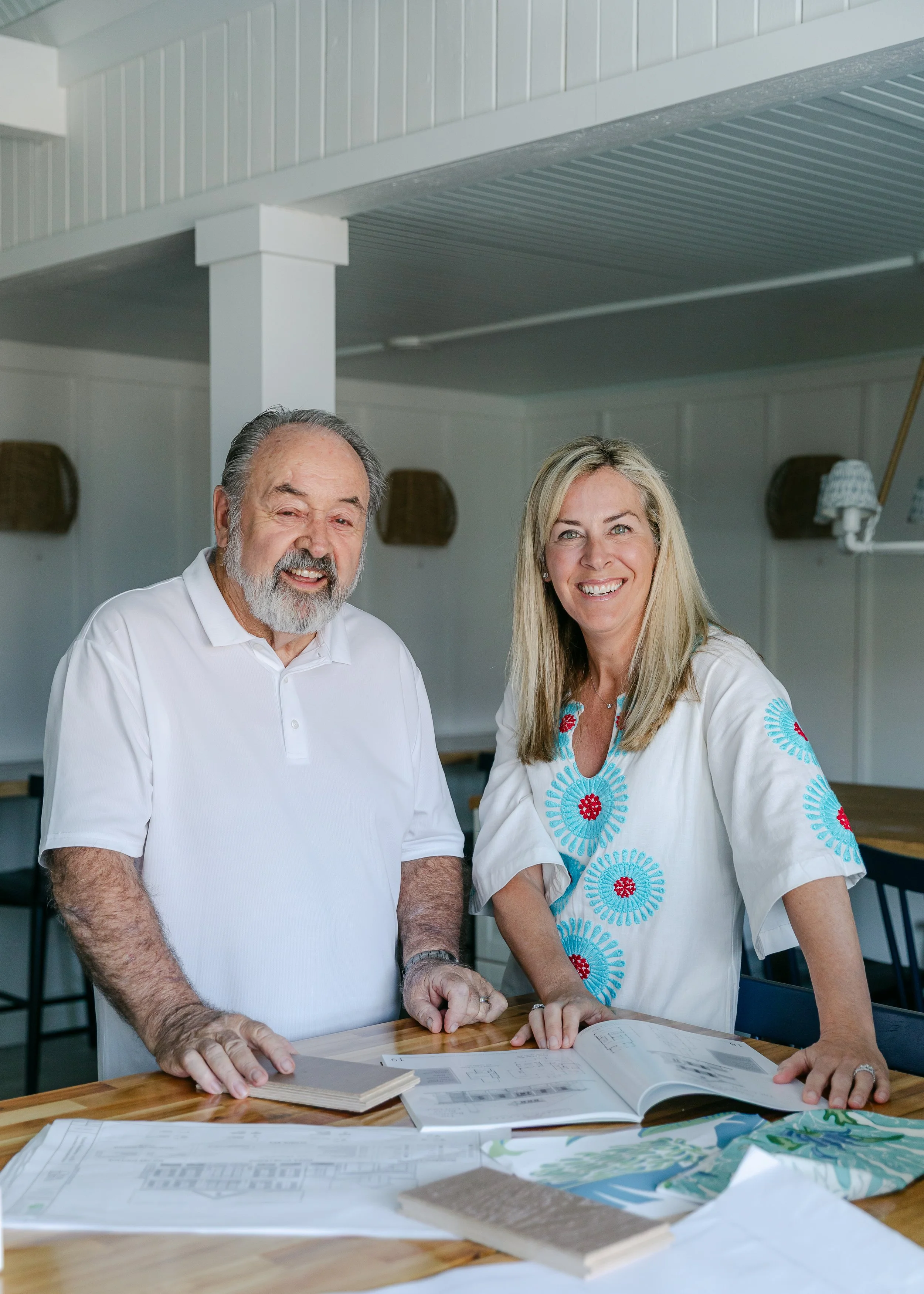Dawn Hillman & Chuck Drace. Cabana Lane Builders at Crane Island. A smiling man and woman standing at a table with architectural plans and books, in a room with wood paneling and wicker baskets on the wall.