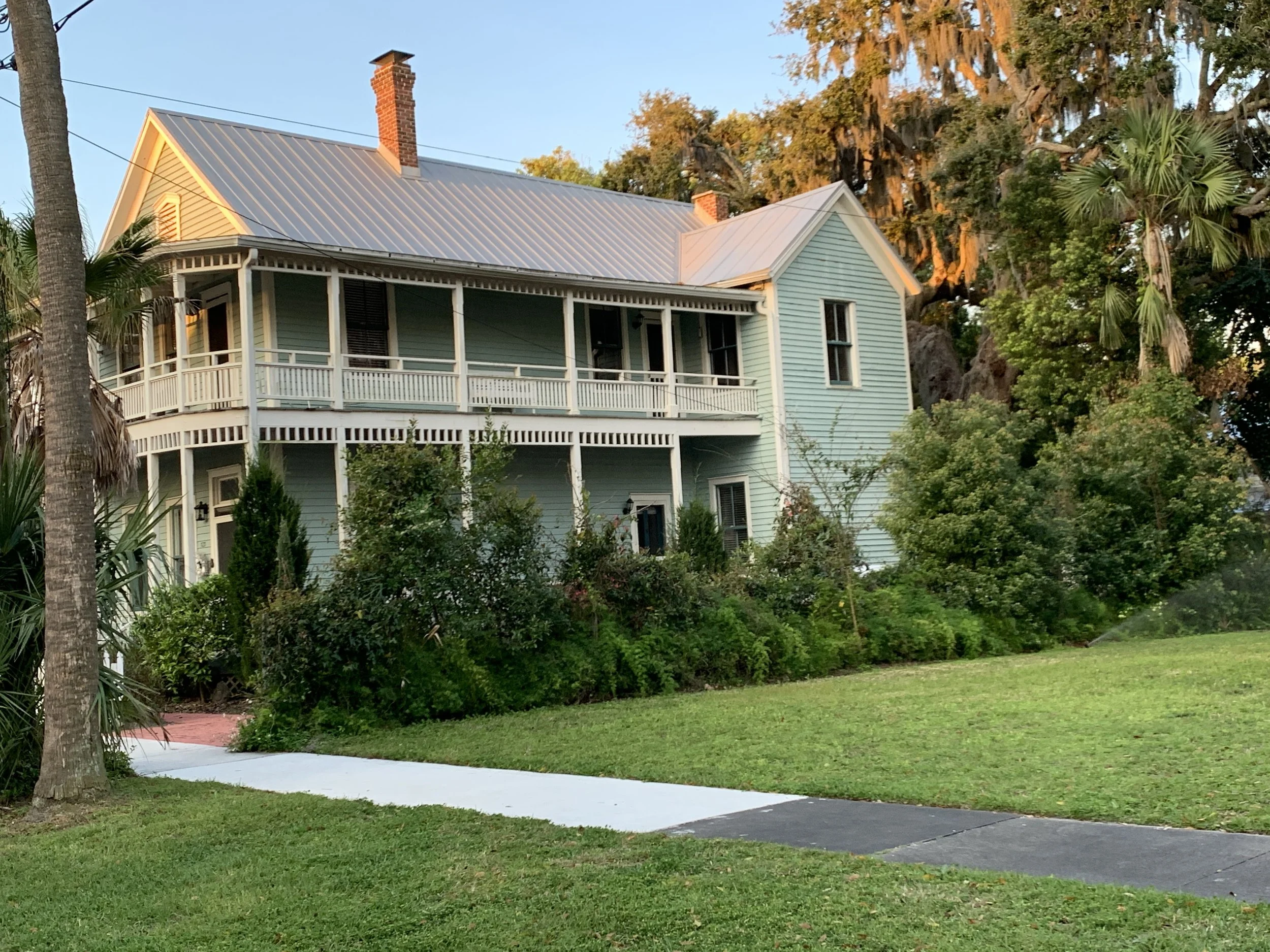 A large, two-story, Victorian-style house painted in light blue with white trim, surrounded by lush green grass and trees, during sunset. Downtown Fernandina Beach, FL - Examples of Lowcountry Architecture.
