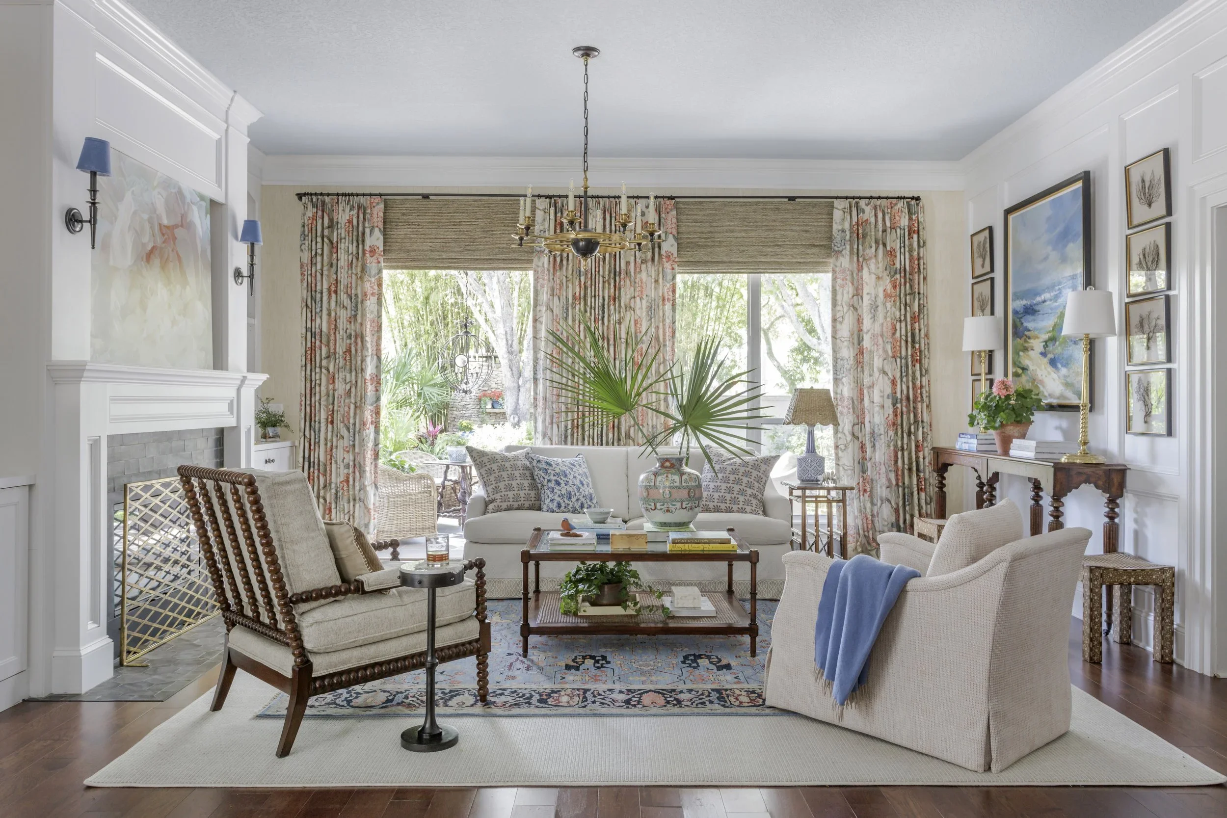 Living room with large windows, floral curtains, white sofa, beige armchairs, wooden coffee table with green plant, patterned rug, side tables with lamps, chandelier, art on walls, and outdoor view with trees.
