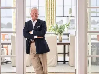 Jim Strickland - Historical Concepts Founder. A smiling man in a navy blazer and beige pants standing with arms crossed in a bright, modern room with large glass windows and a view of a balcony or patio.
