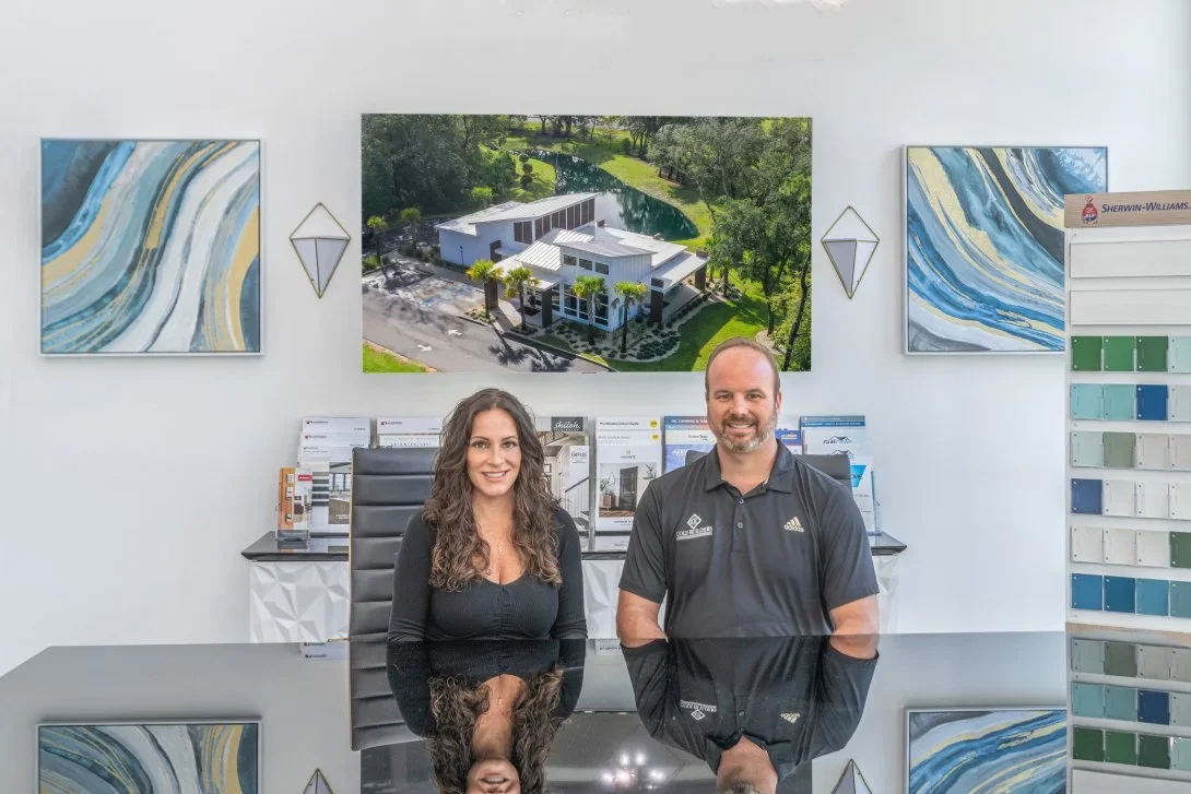 A man and a woman sitting at a glossy black table in an office showroom, with three abstract paintings on the white walls behind them, a large aerial photo of a modern building and landscape above them, and a display of paint color samples on the rig