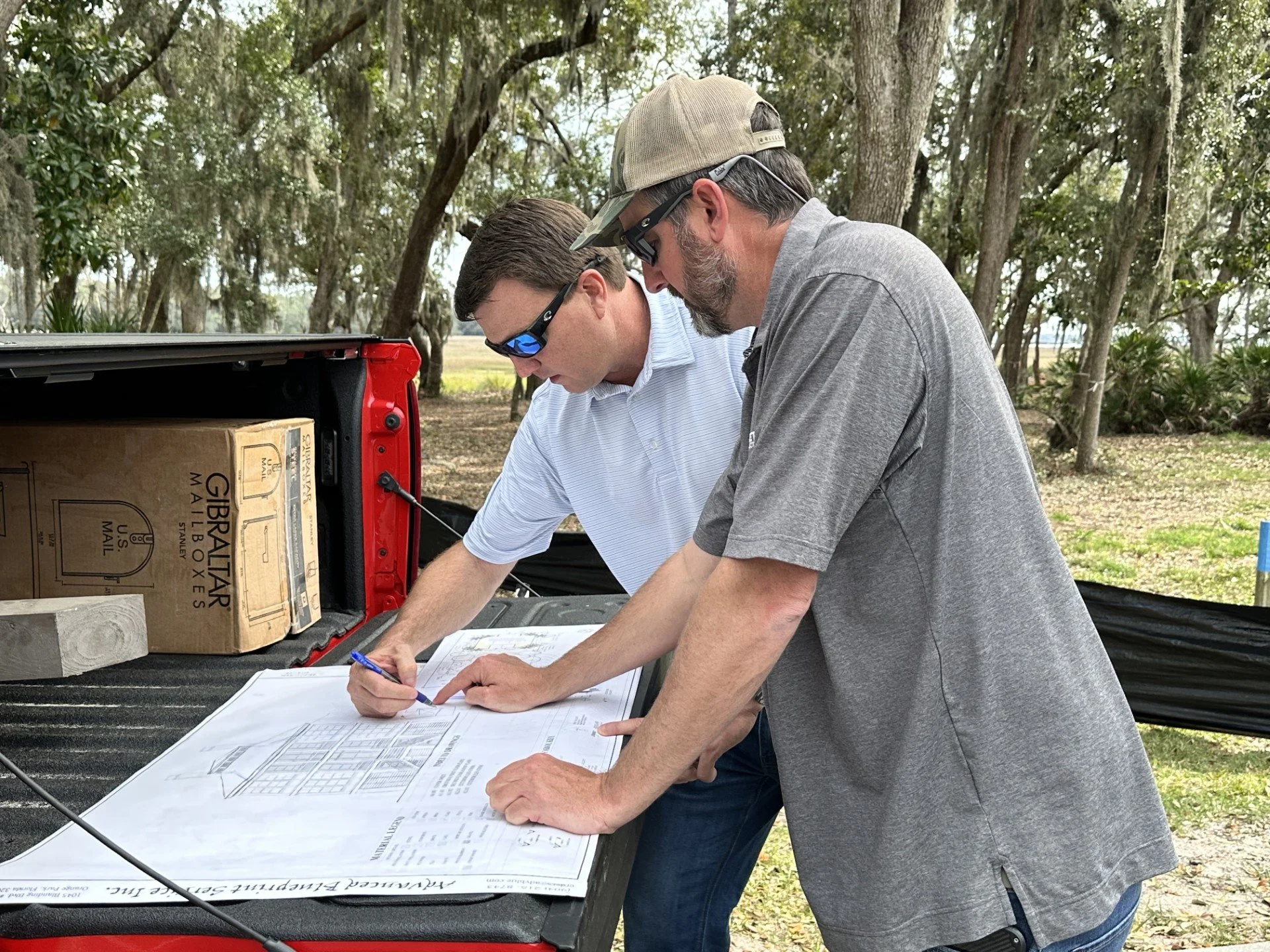 Two men reviewing blueprints outdoors next to an open truck bed with a cardboard box in a wooded area.