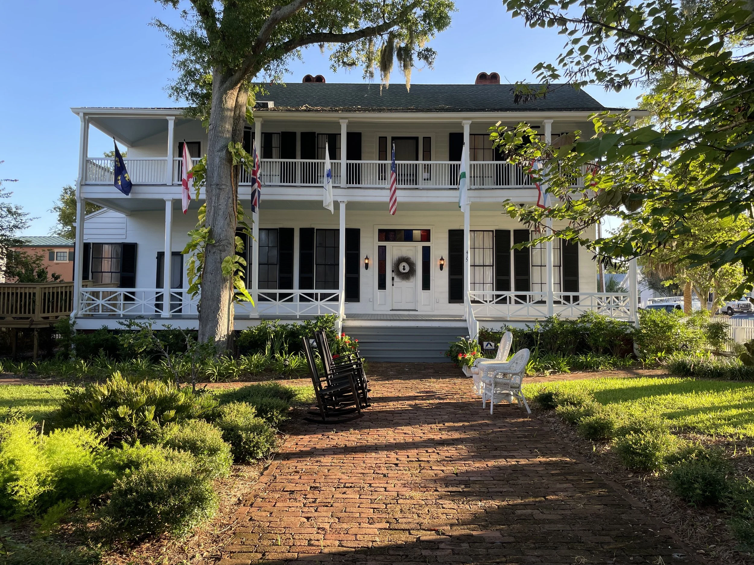 A large two-story white house with black shutters, a front porch, and a staircase leading to the entrance. Flags are displayed on poles along the porch. There is a brick pathway lined with small bushes and plants, leading to the house. Several chairs