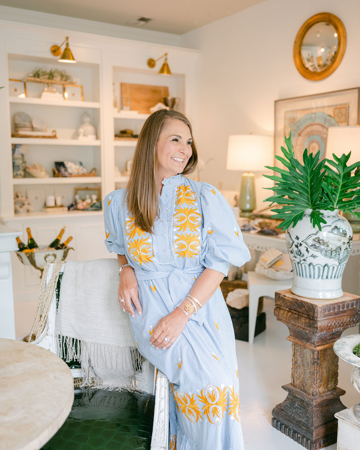 Leah Bailey Interior Design - Crane Island. A woman in a blue and yellow embroidered dress smiling in a decorated living room with shelves, lamps, and a large potted plant.
