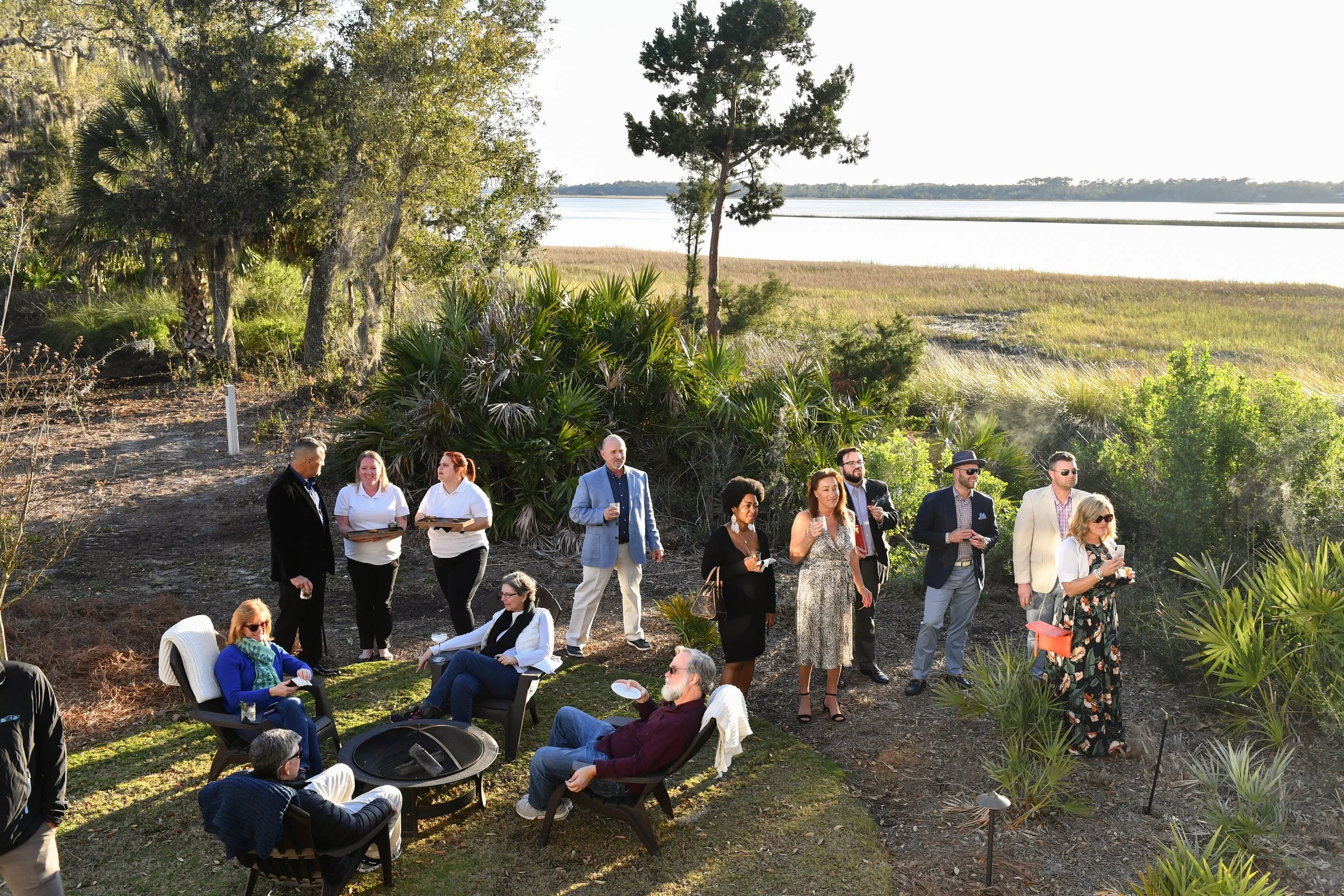 2019 Southern Living Idea House. Group of people at an outdoor gathering beside a lake with trees and grassy wetlands in the background.