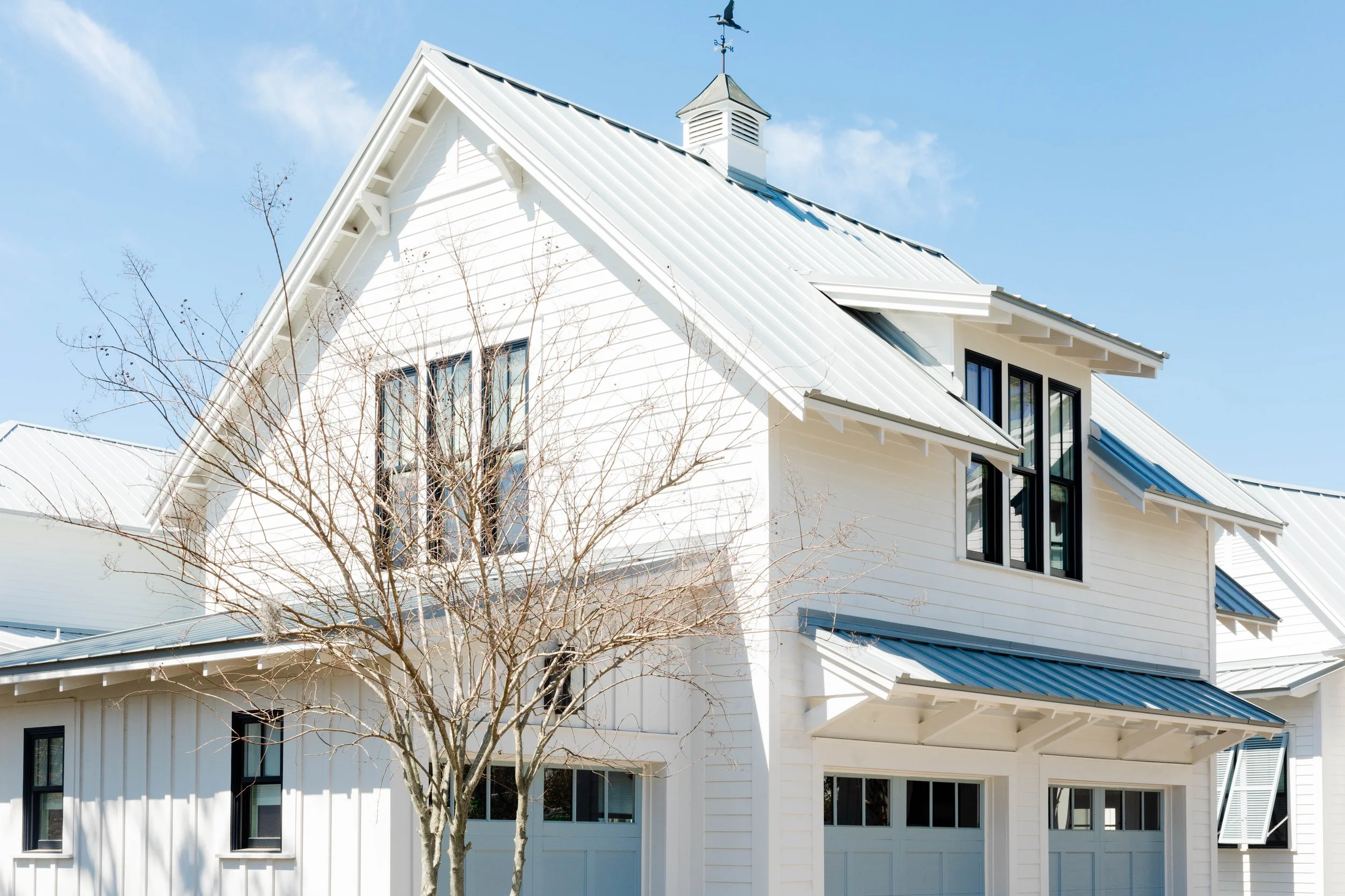 Lake & Land Studio - Crane Island. A white modern farmhouse with a metal roof, large windows, and a small leafless tree in front, under a clear blue sky.