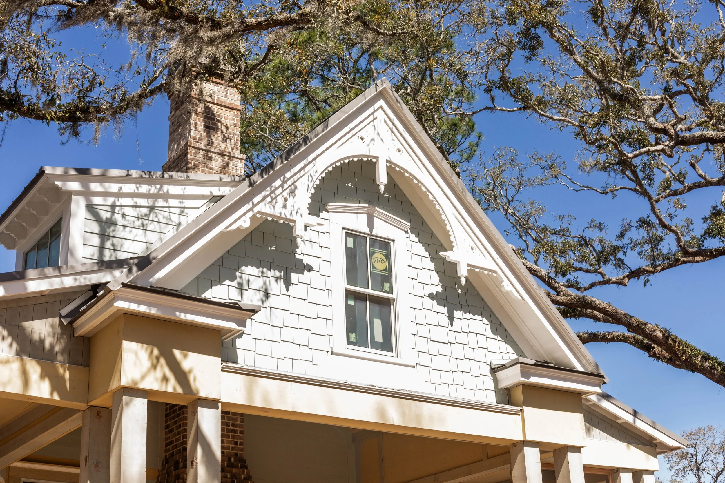 Crane Island Homes. Close-up of a house's gable and window with decorative details, siding, and a brick chimney, under a clear blue sky with tree branches overhead.