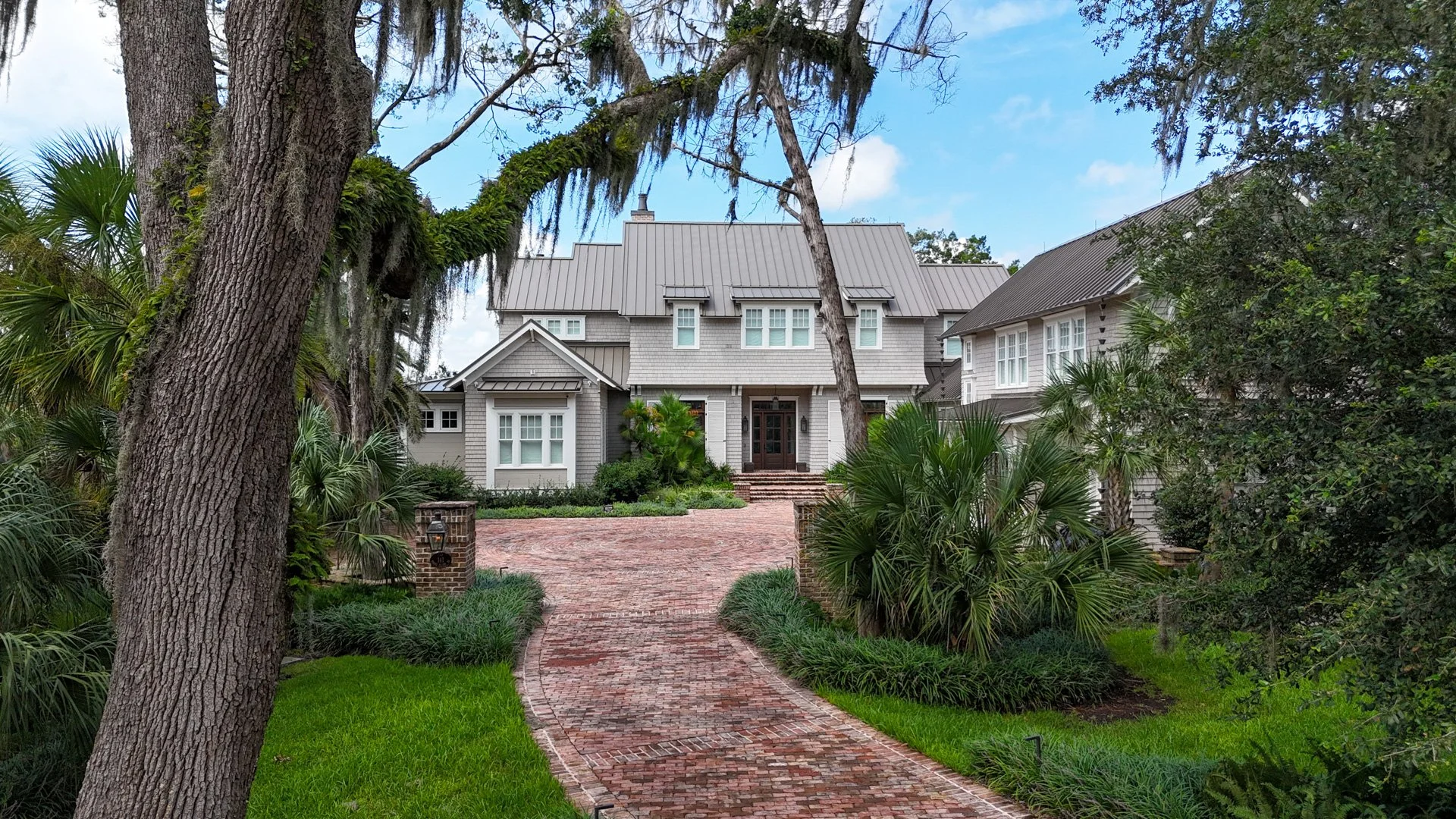 Front view of a large house with a brick pathway leading to the front door, surrounded by lush greenery and trees.