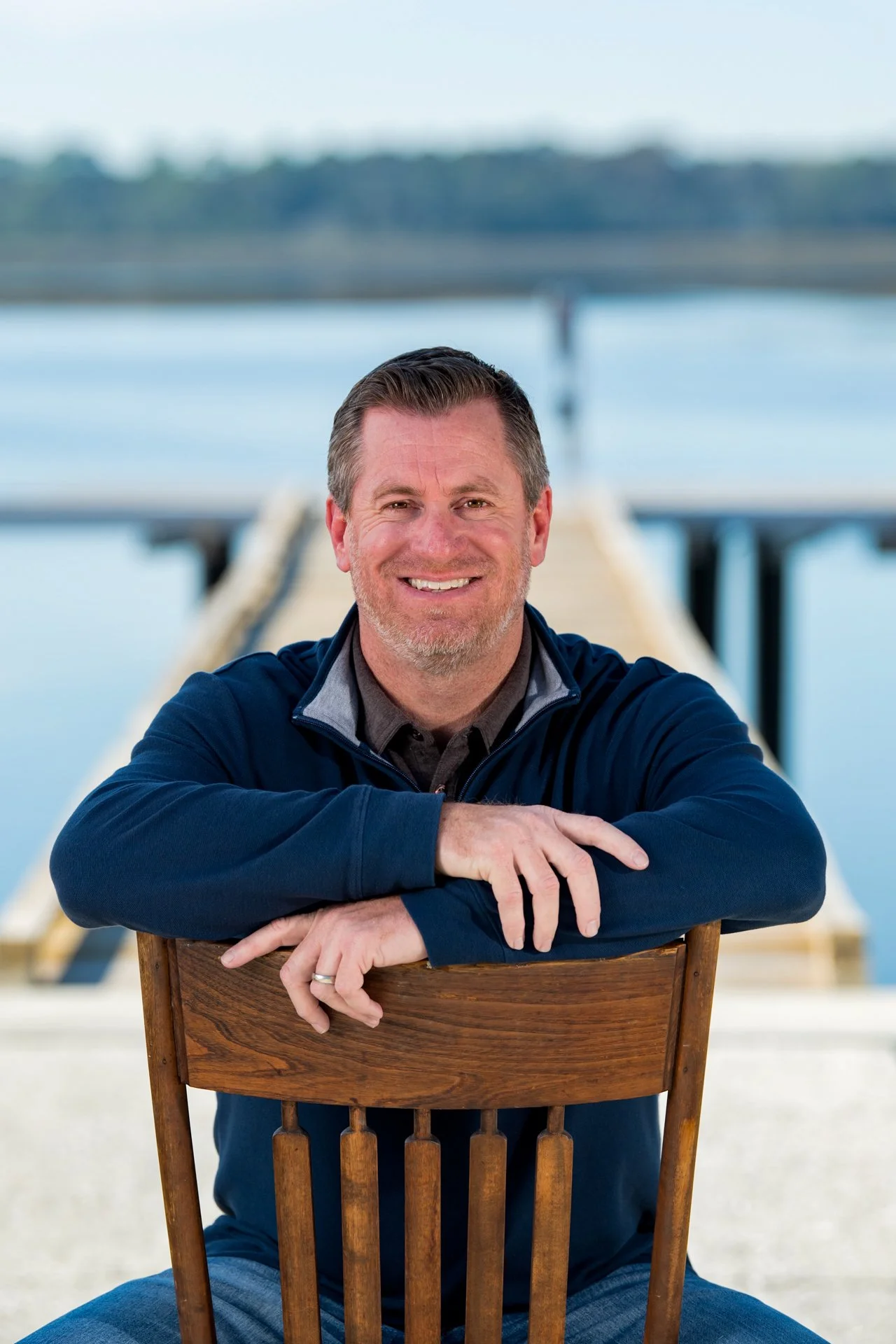 A smiling man sitting on a wooden chair at a dock by a body of water with a bridge in the background.