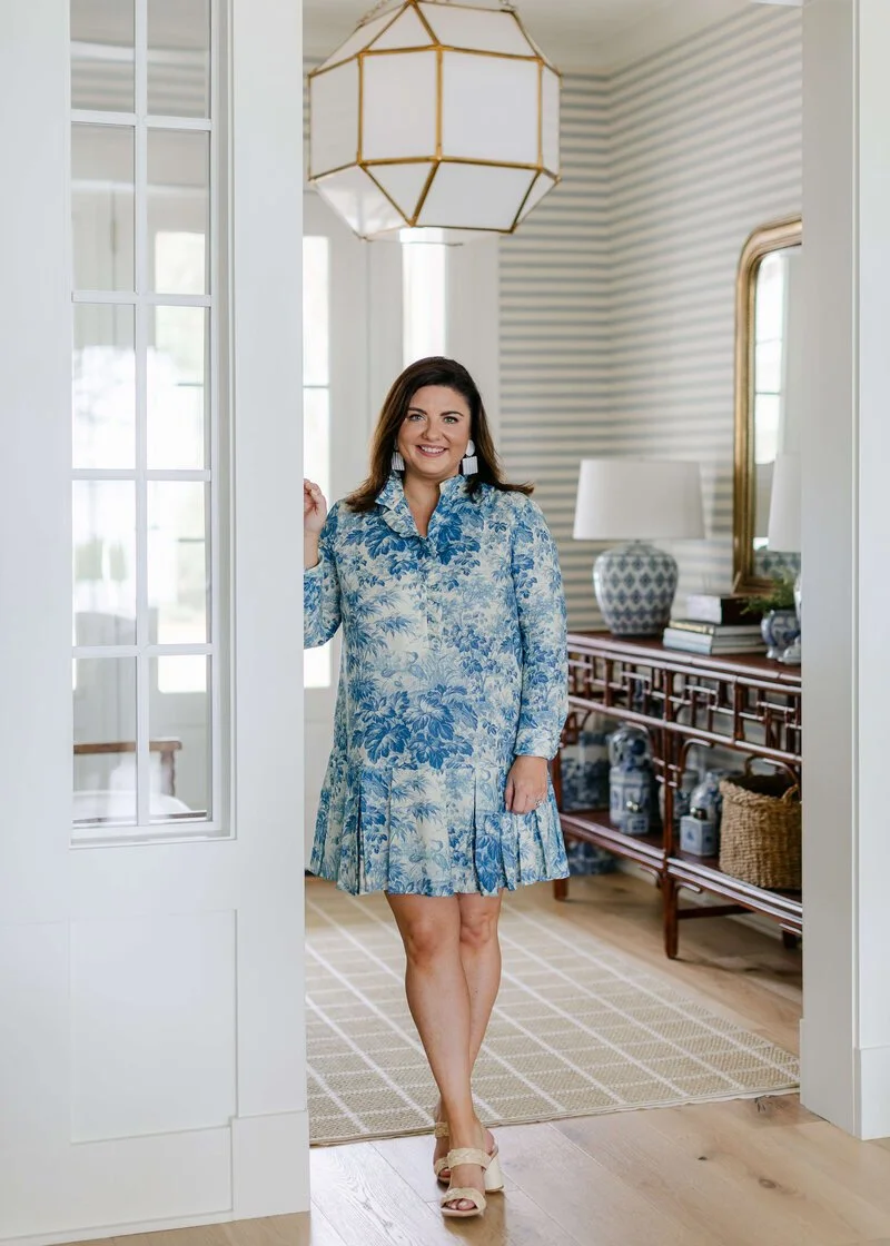 Lola Interiors - Crane Island. A woman in a blue and white floral dress standing in a bright, decorated room with striped wallpaper, a wooden console table, lamps, and blue and white vases.