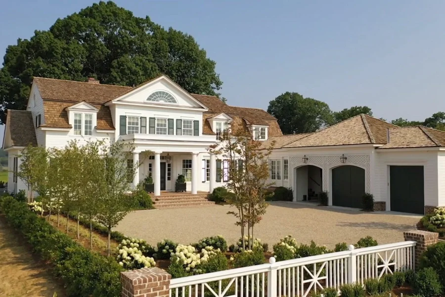 Large, elegant white house with a brown shingled roof, surrounded by well-kept landscaping and a white picket fence.