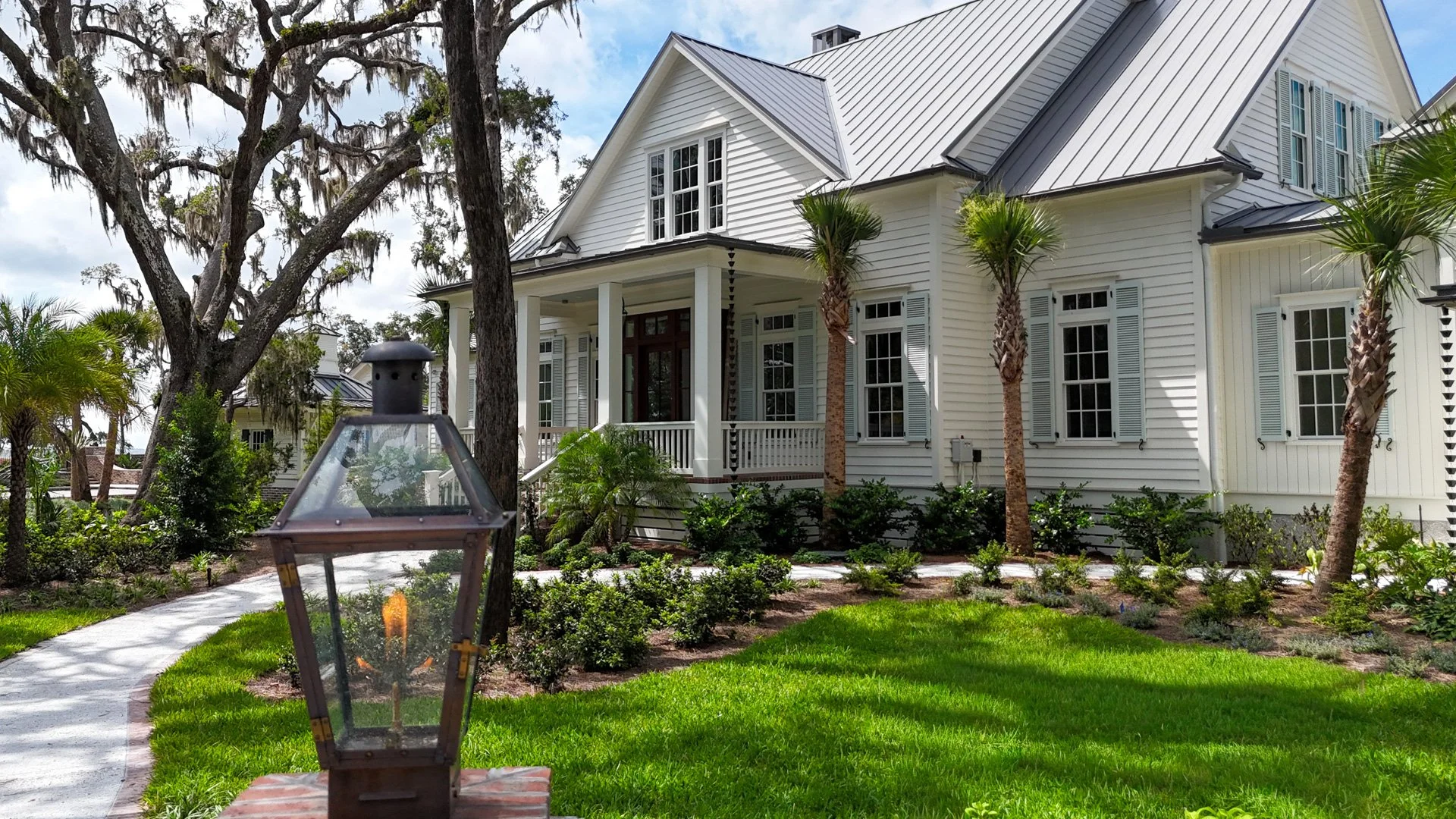 Hansen Savannah, Ga. Home at Crane Island. A large white house with a metal roof, multiple windows with shutters, and a front porch, surrounded by a landscaped yard with grass, bushes, and palm trees, under a partly cloudy sky.