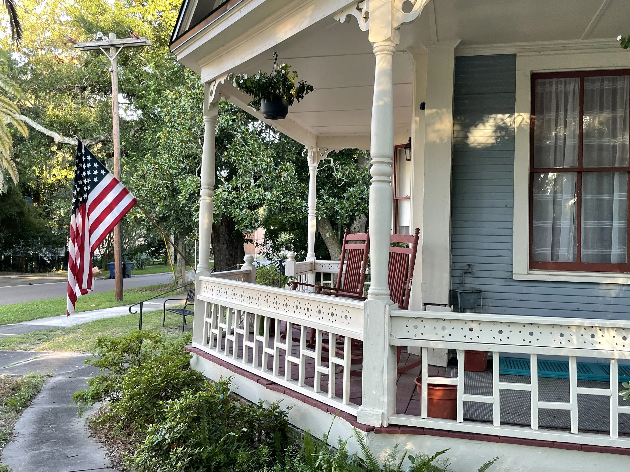 Front porch of a house with white railings, two red rocking chairs, an American flag, and lush green trees in the background.Downtown Fernandina Beach, FL - Examples of Lowcountry Architecture.