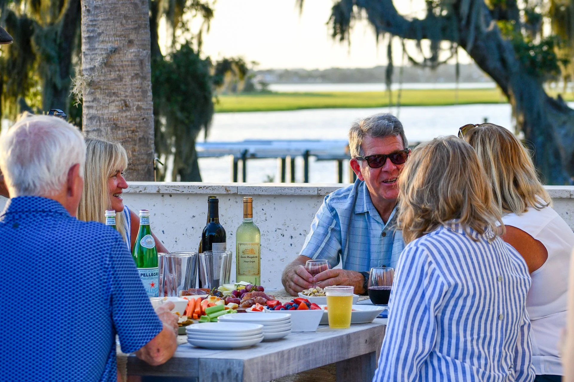 Lifestyle at Crane Island ,FL . A group of people enjoying a meal outdoors by a lake, with a table filled with drinks, fruits, vegetables, and snacks, surrounded by trees.