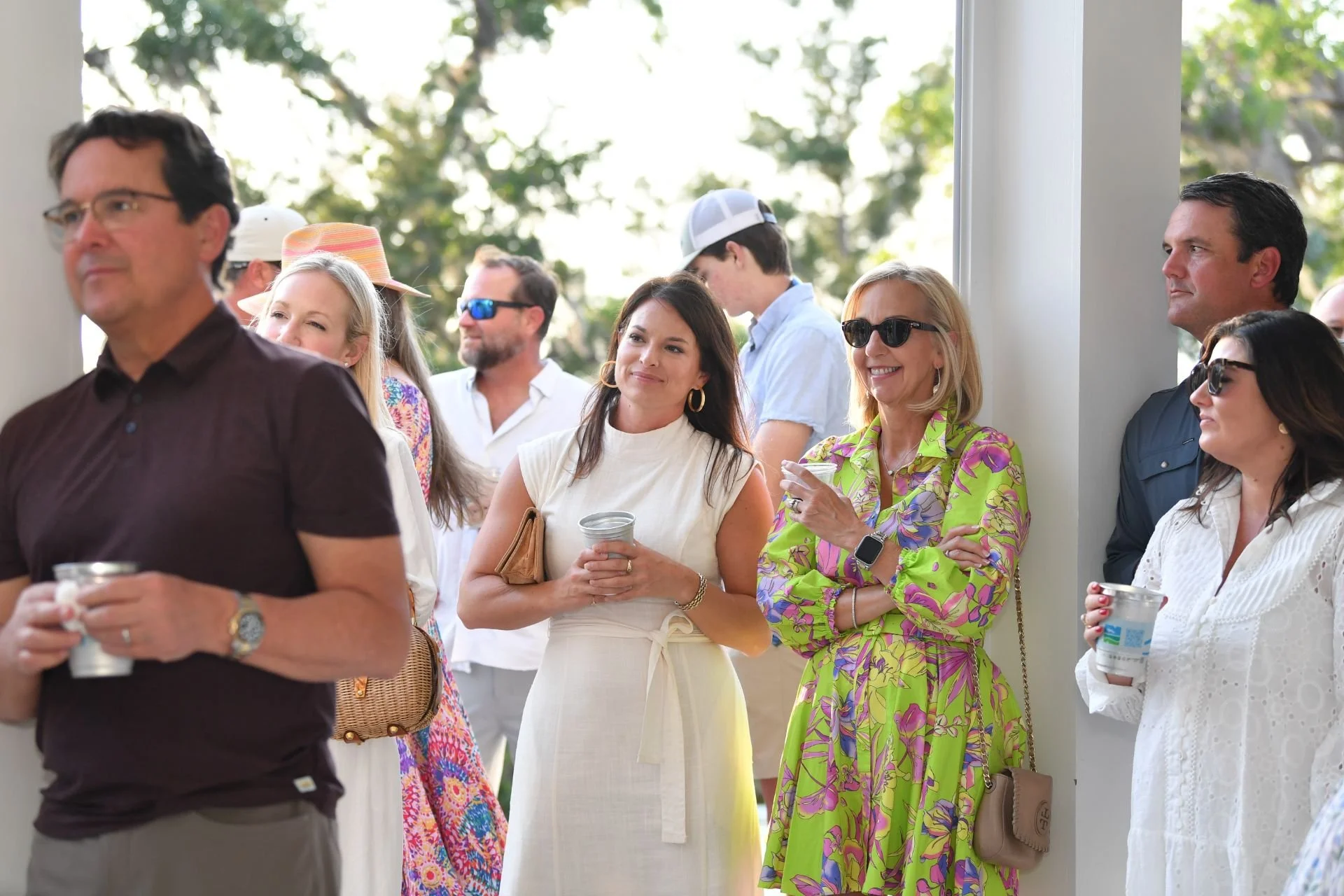 Crane Island Event. Group of people attending an outdoor social event, some wearing sunglasses and holding drinks, standing near a white column with trees in the background.