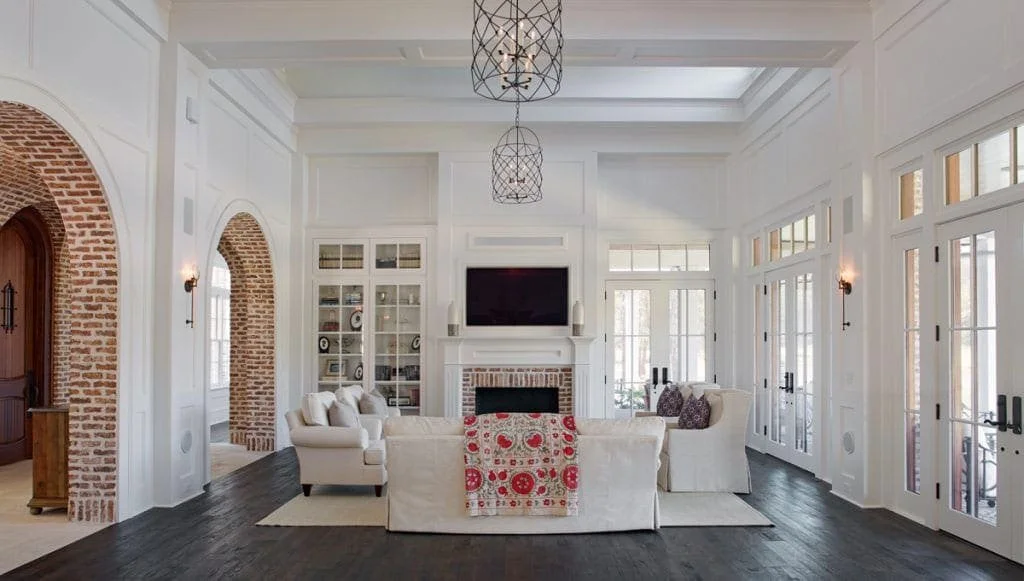 Bright living room with white walls and ceiling, dark hardwood floors, large windows and glass doors, white furniture, a fireplace, and a mounted television.