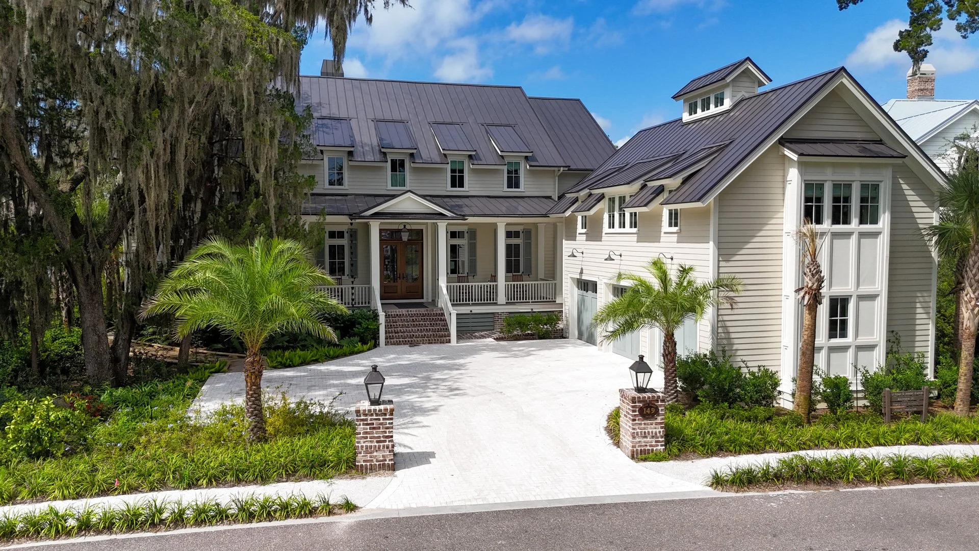 Pearce Scott Architects - Crane Island. A large two-story house with white siding, black metal roof, and front porch, surrounded by palm trees and greenery, with a paved driveway and brick pillars with lamps at the entrance.