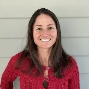Lauren Kelly - Moser Kelly Design Group - Crane Island. A woman with long dark hair smiling, wearing a red knit sweater, standing in front of a light-colored wall.