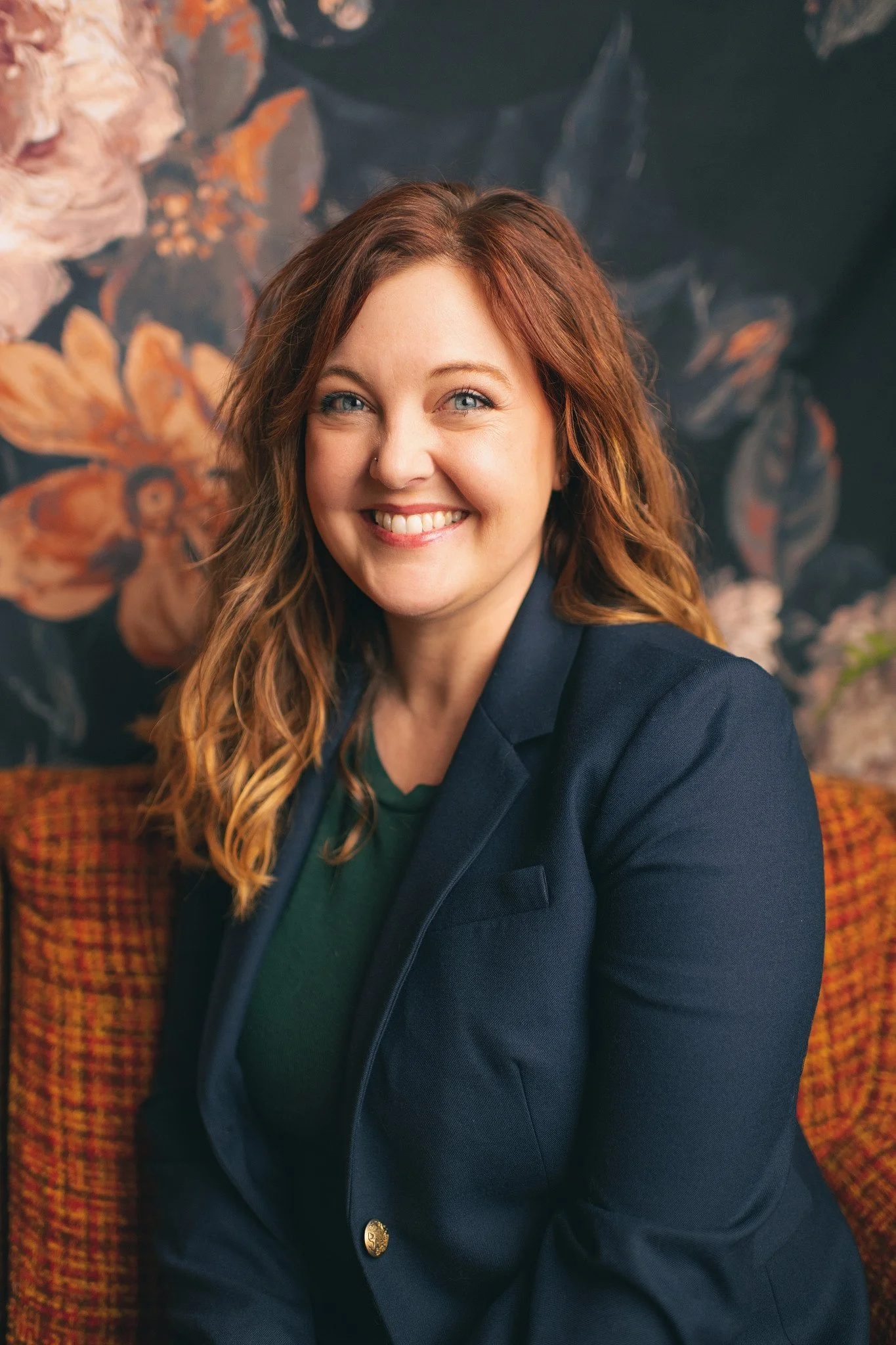 A woman with wavy red hair smiling, wearing a navy blazer and green top, sitting on a patterned orange and brown chair against a floral background.