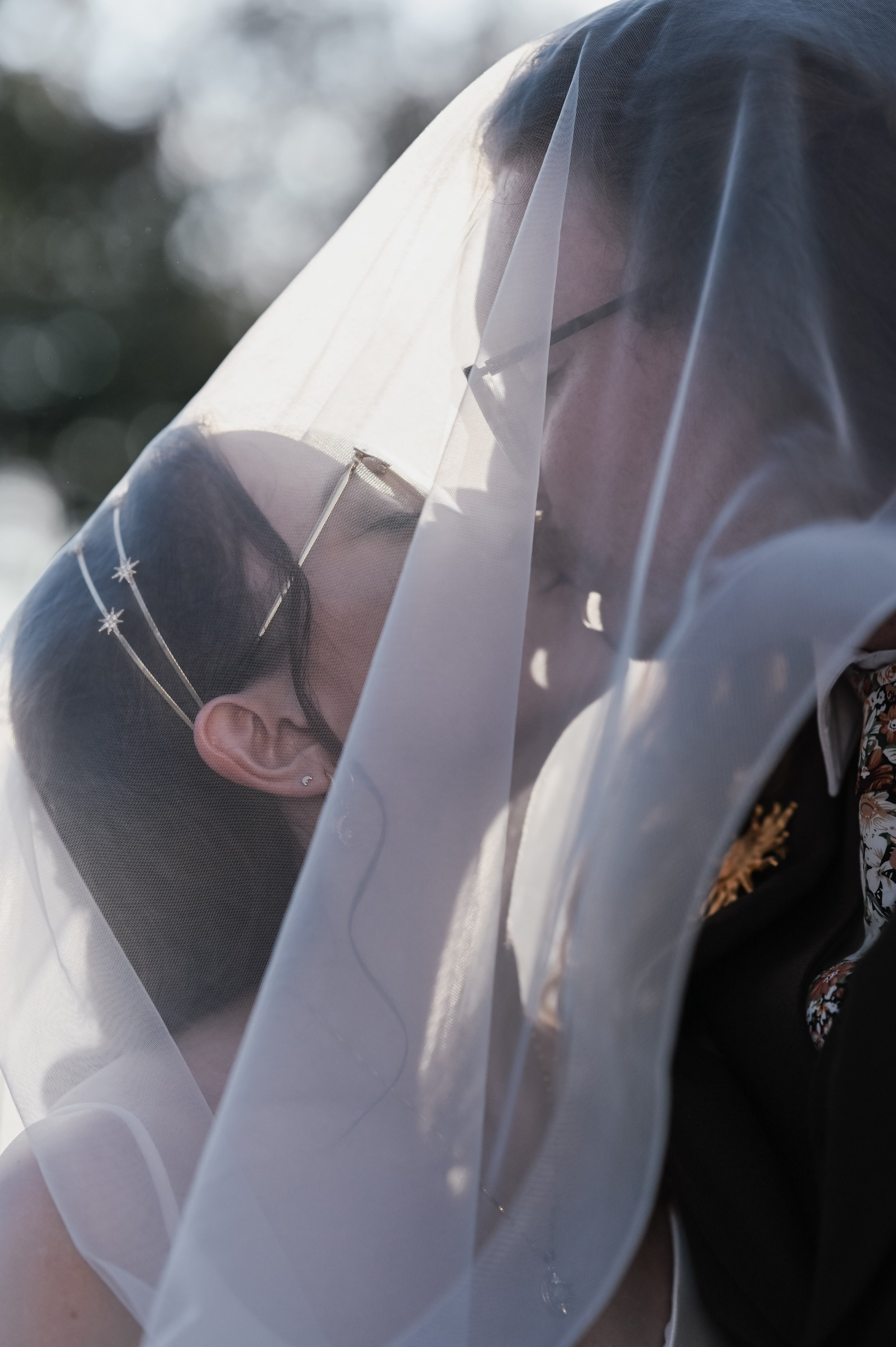 A bride and groom sharing a kiss through a sheer, white veil outdoors.