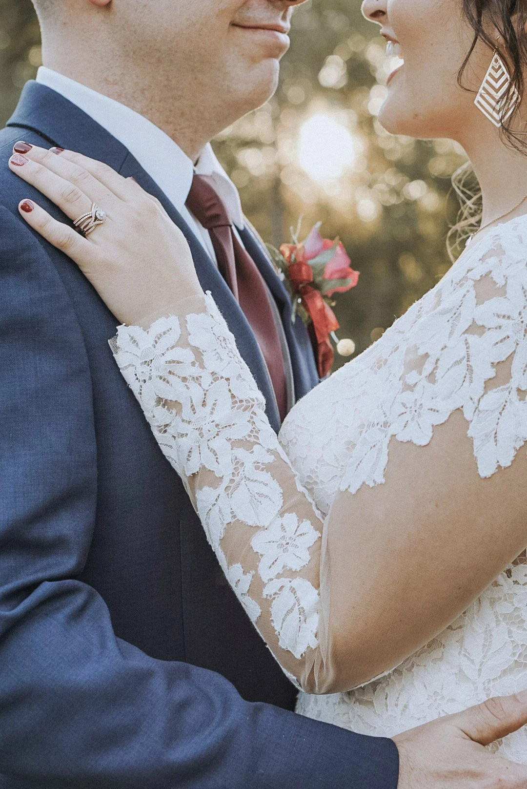 A bride and groom embrace outdoors, focusing on their faces and upper bodies. The bride wears a white lace dress with floral patterns and has lace sleeves, her hand resting on the groom's shoulder. The groom is dressed in a blue suit with a white shi