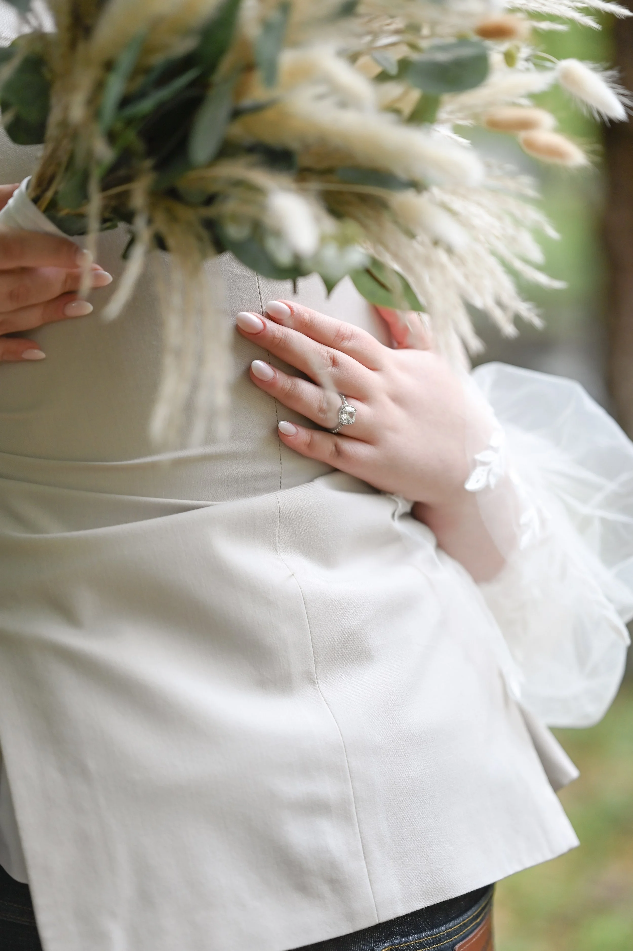 Close-up of a bride's hand with an engagement ring resting on her wedding dress. She holds a bouquet of flowers and greenery. The background is blurred.