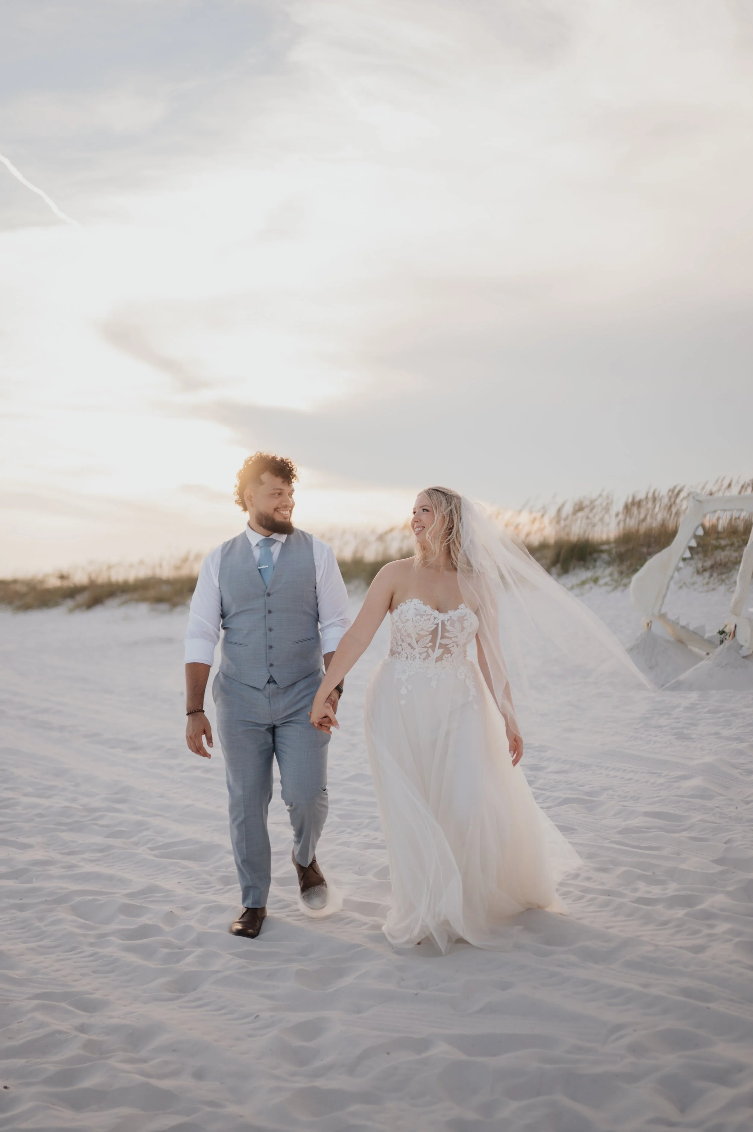 A newlywed couple walking hand in hand on a sandy beach during sunset, dressed in wedding attire with the bride in a lace wedding gown and the groom in a gray suit.