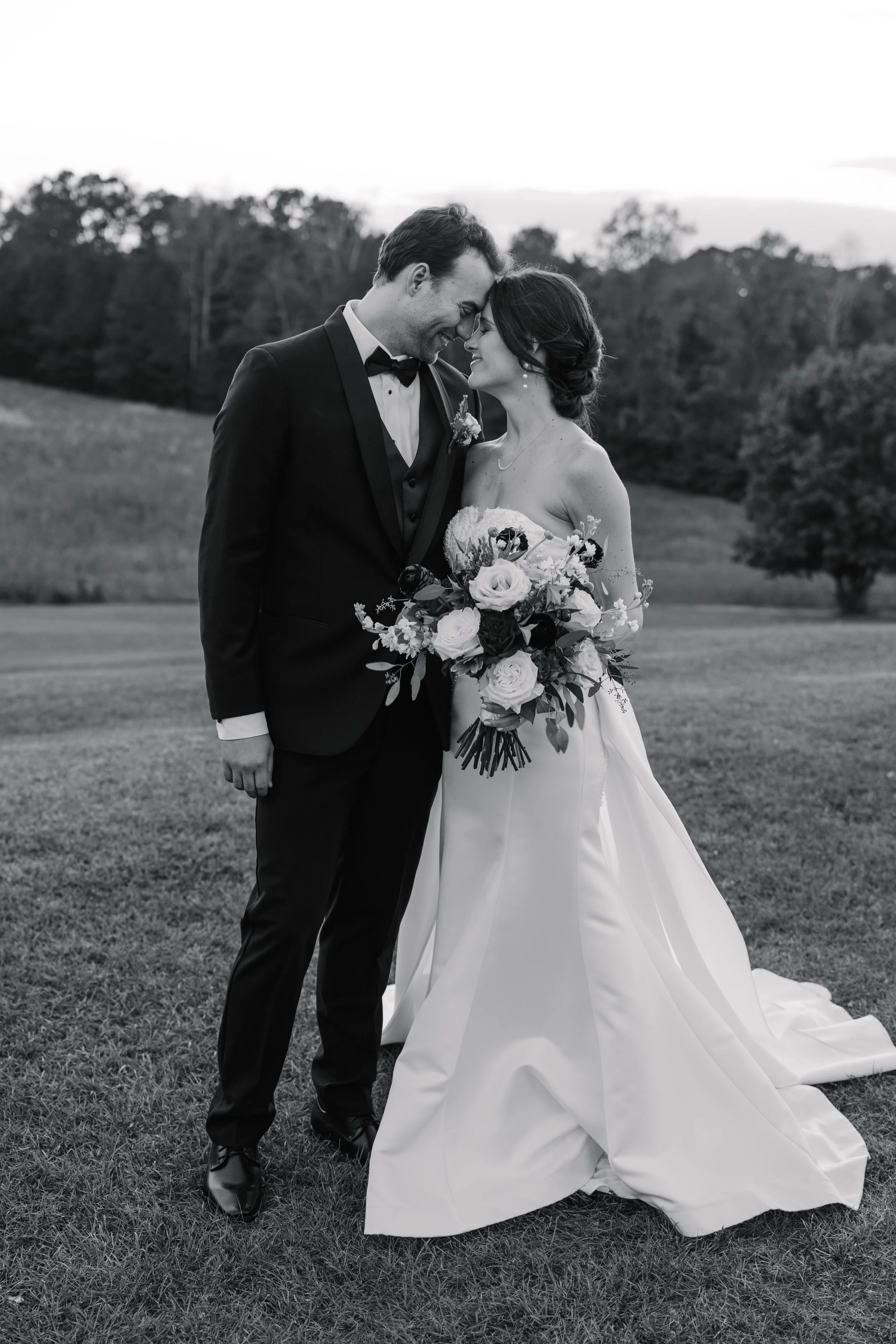 Black and white photo of a bride and groom standing closely with foreheads touching, smiling. The groom wears a tuxedo, and the bride wears a strapless wedding gown and holds a bouquet of flowers. They are outdoors on a grassy field with trees in the