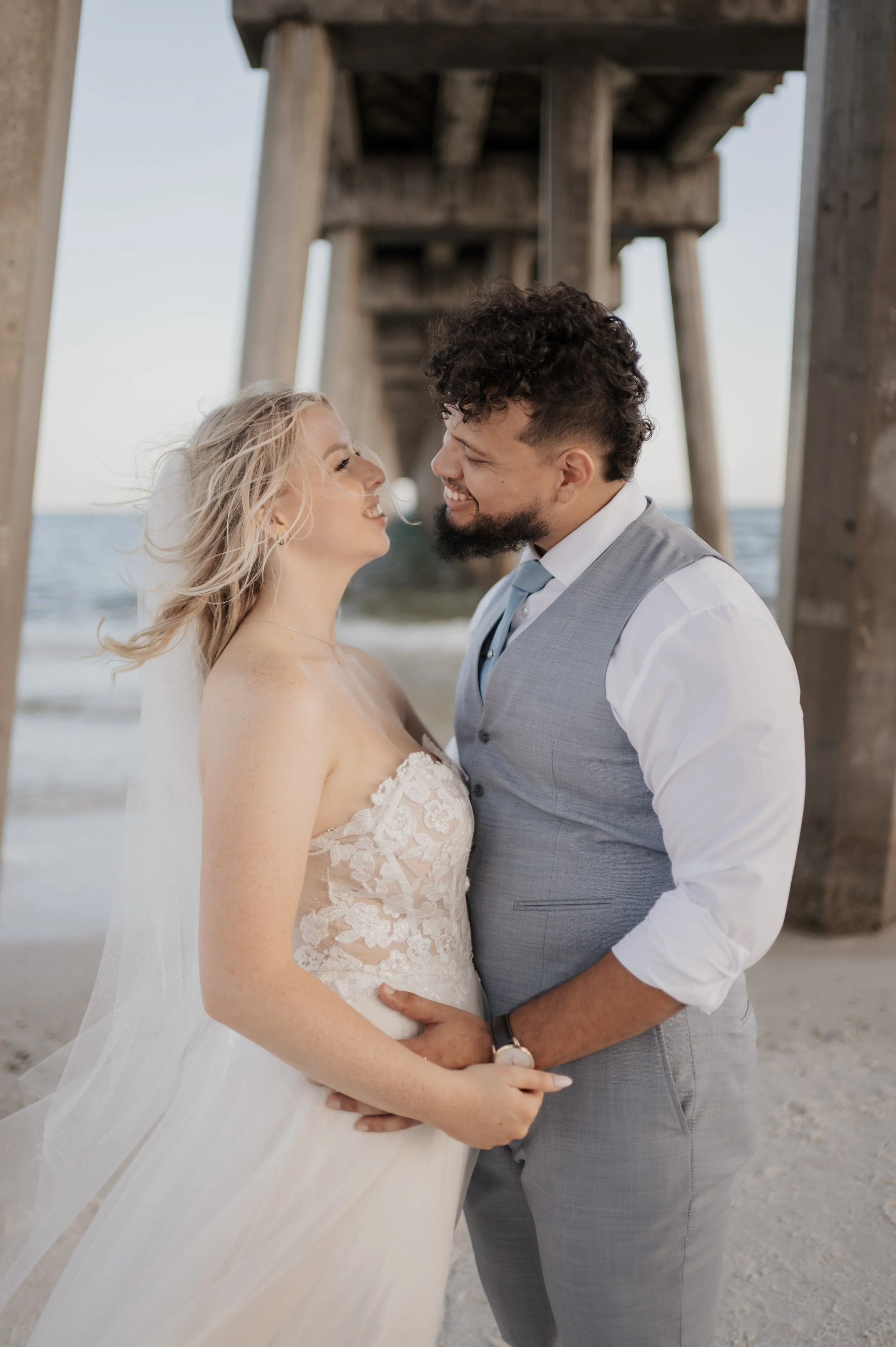 A happy bride and groom on the beach, close together under a pier, smiling at each other during their wedding.