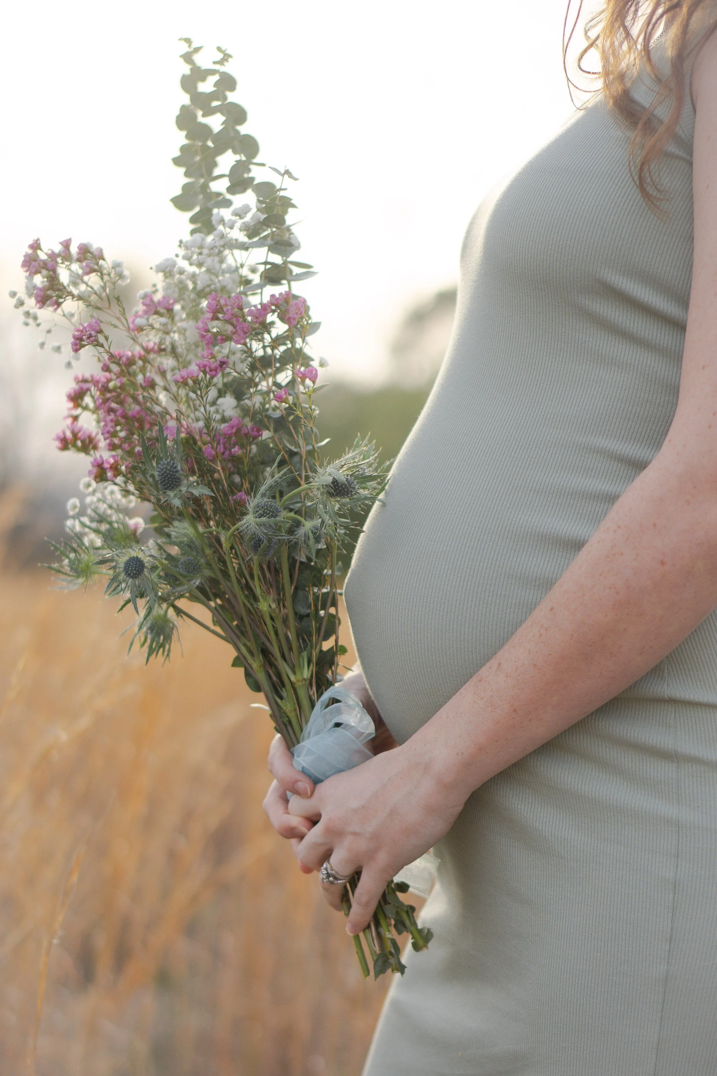 Close-up of a pregnant woman holding a bouquet of wildflowers in an outdoor setting with golden hour sunlight.