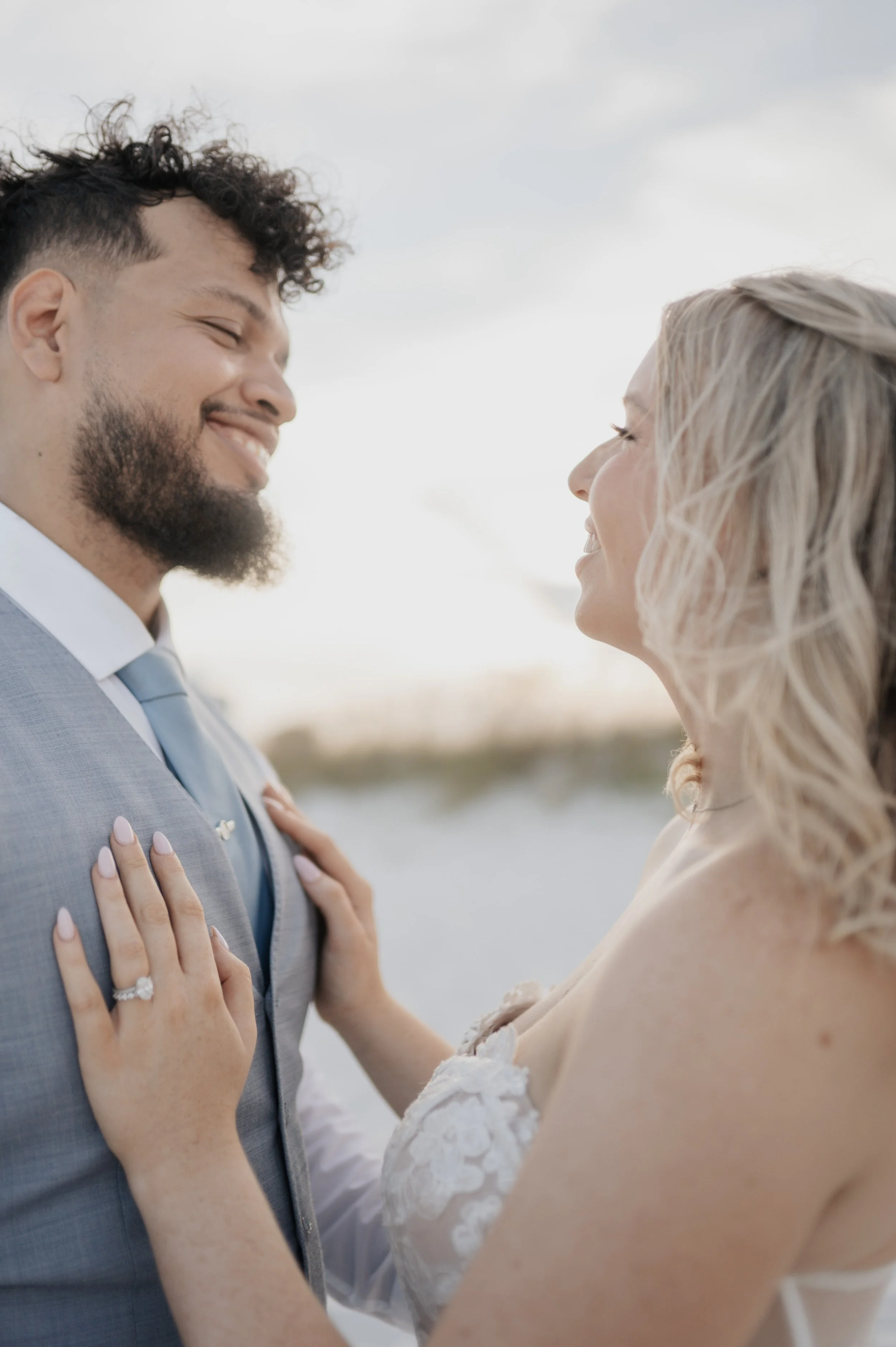 A smiling couple in wedding attire standing close together outdoors, with water and an overcast sky in the background.