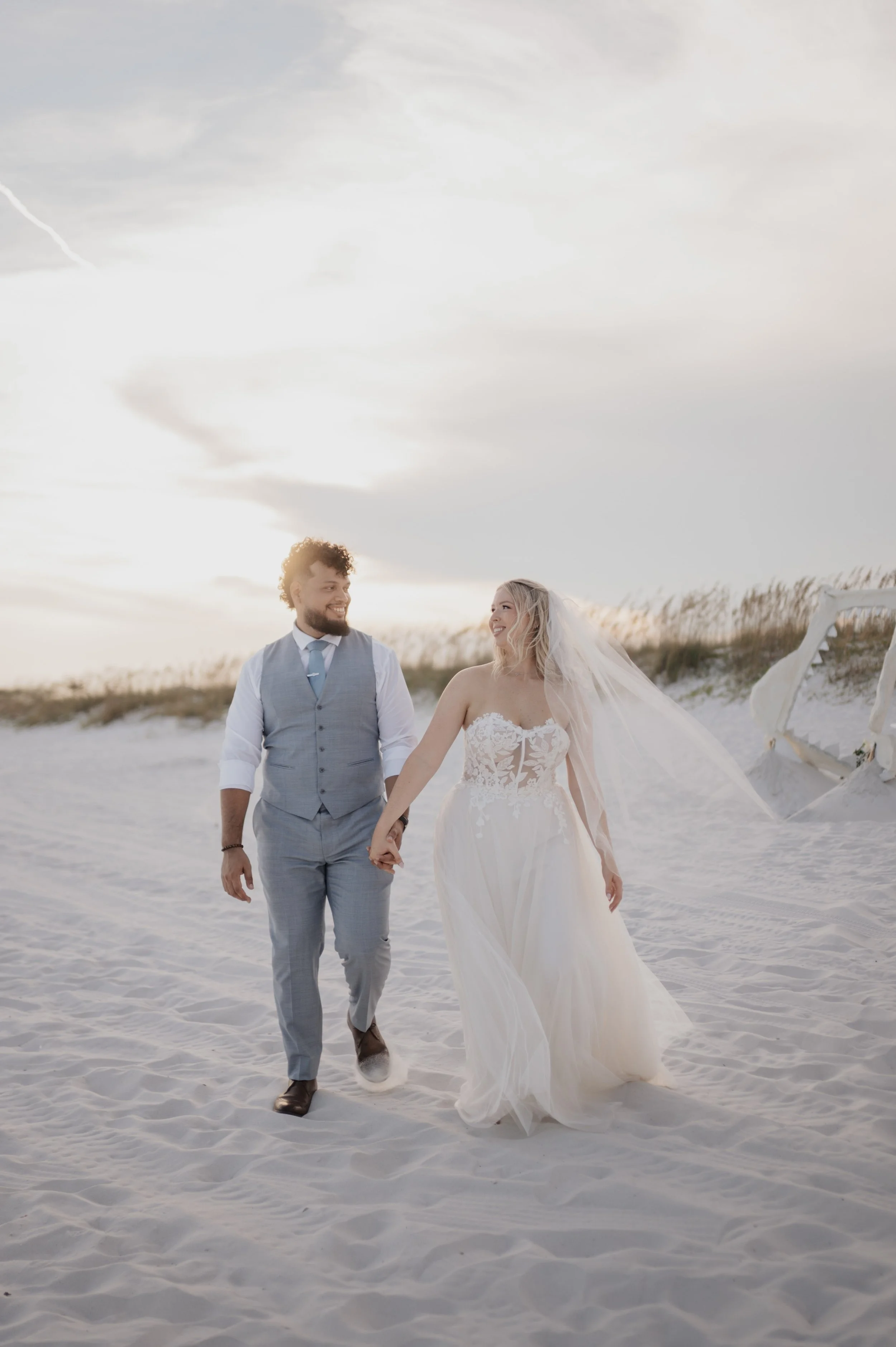 A couple in wedding attire walking hand-in-hand on a sandy beach during sunset.