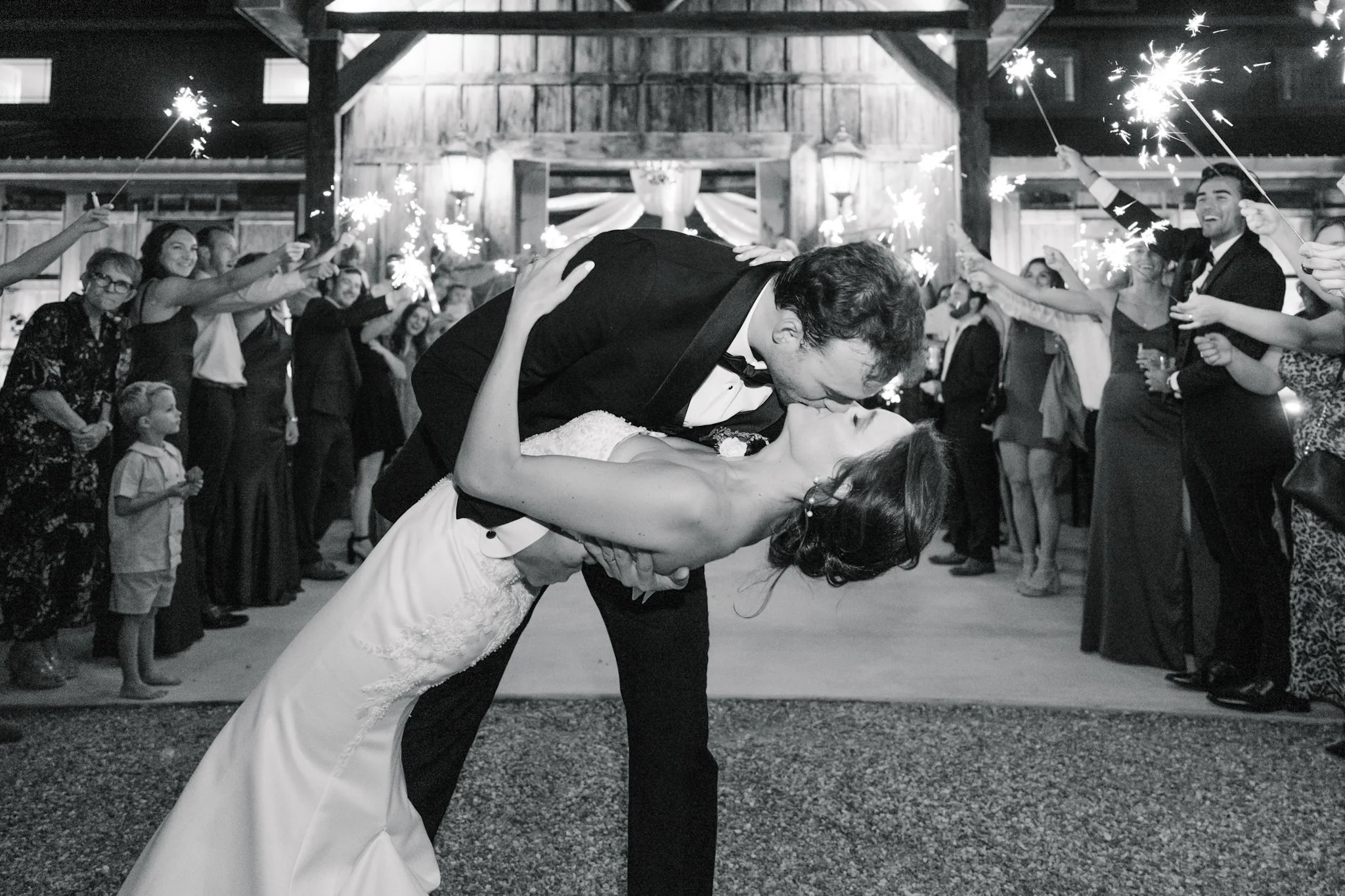 A newlywed couple shares a kiss during their wedding reception, with guests holding sparklers in the background.