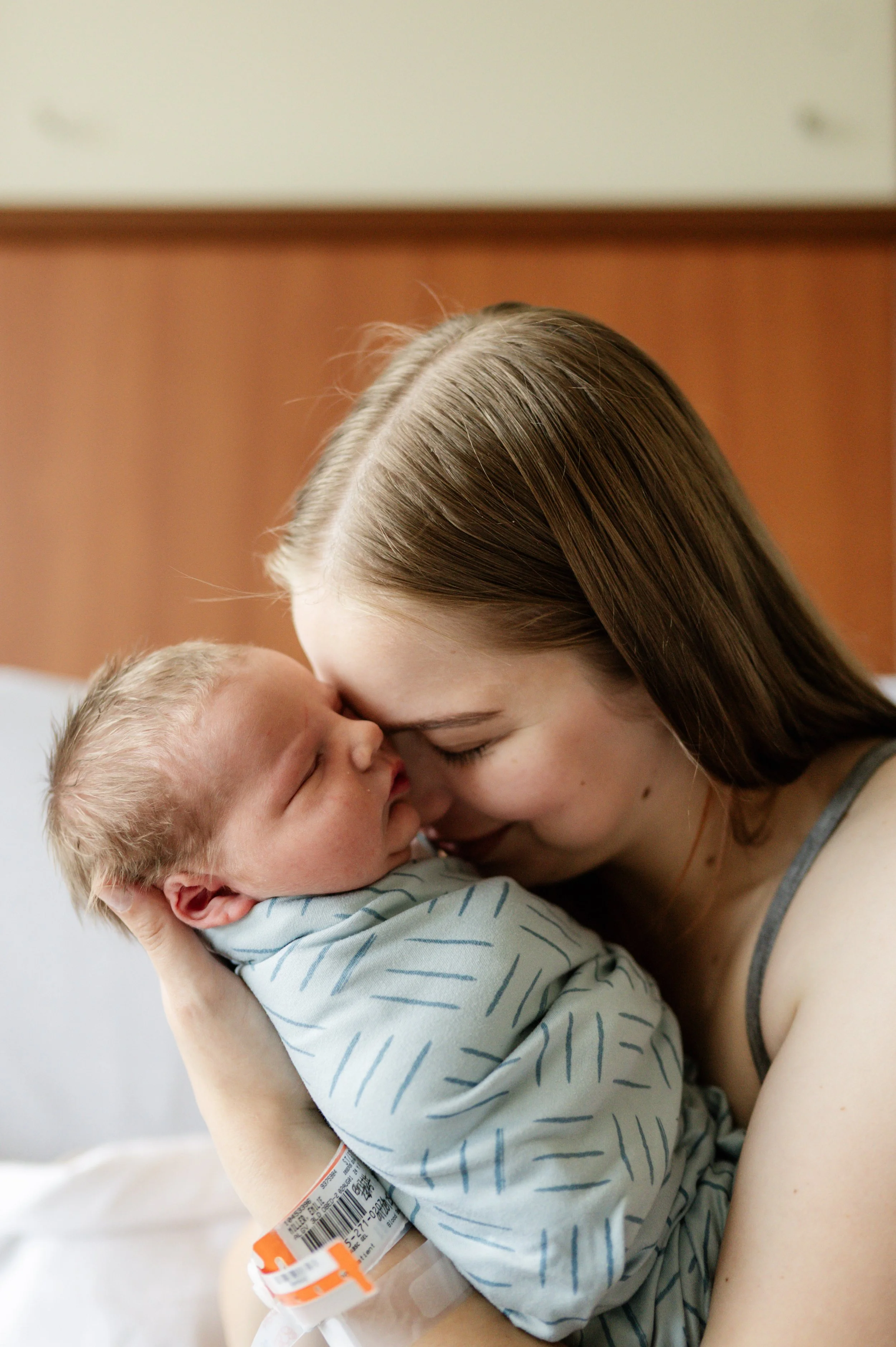 Young woman holding a newborn baby in a hospital, both with foreheads touching, sharing a tender moment.