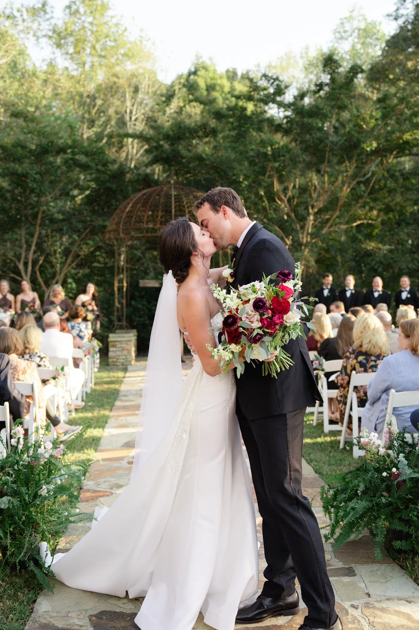 A bride and groom sharing a kiss during their outdoor wedding ceremony, with guests seated in the background and lush greenery surrounding them.