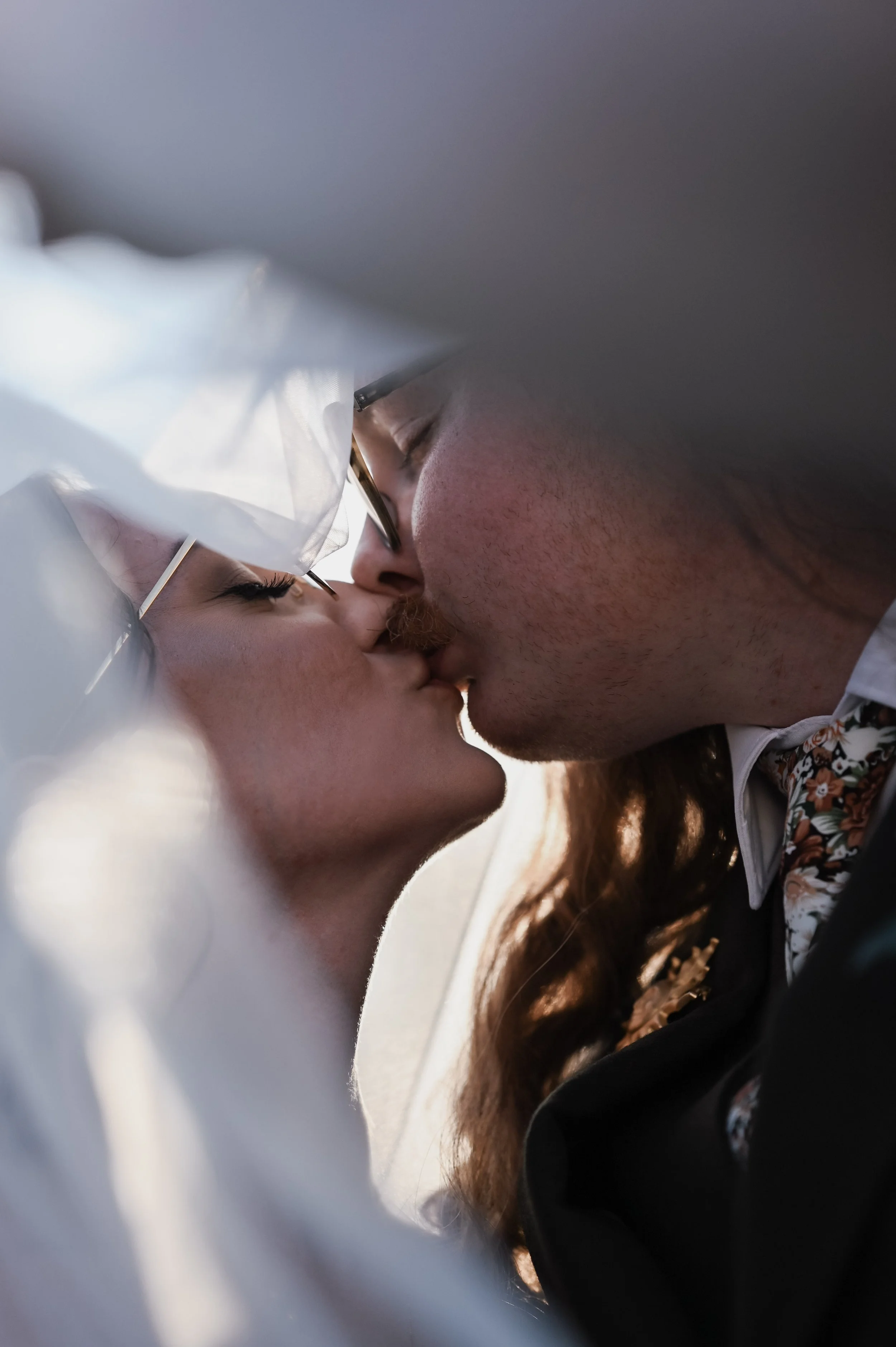 A close-up of a couple sharing a kiss, with both wearing glasses and fine clothing, possibly attending a formal event.