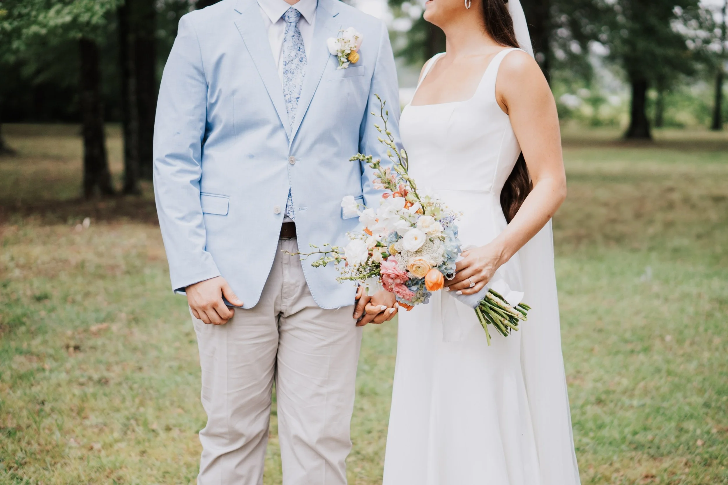 A bride and groom stand outdoors in a grassy area, holding hands. The bride wears a white wedding dress and holds a colorful bouquet of flowers. The groom wears a light blue blazer, a floral tie, and beige pants.