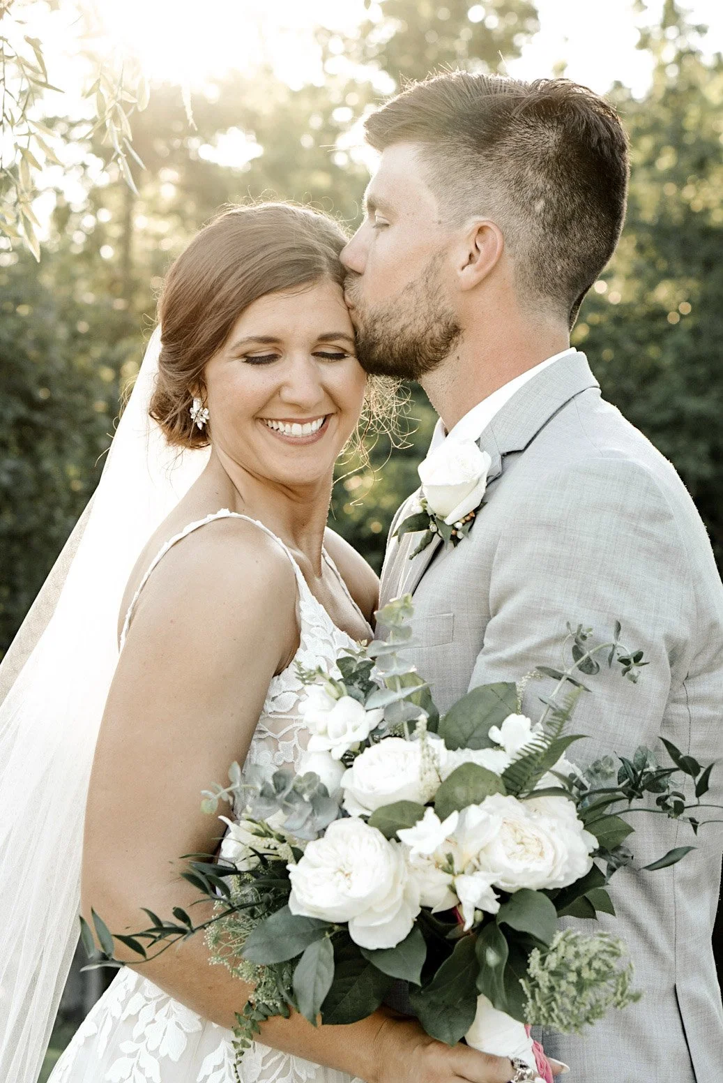 A newlywed couple on their wedding day outdoors, with the groom kissing the bride's temple. The bride holds a bouquet of white roses and greenery, wearing a lace wedding dress. The groom wears a light gray suit with a white shirt and boutonniere. Sun