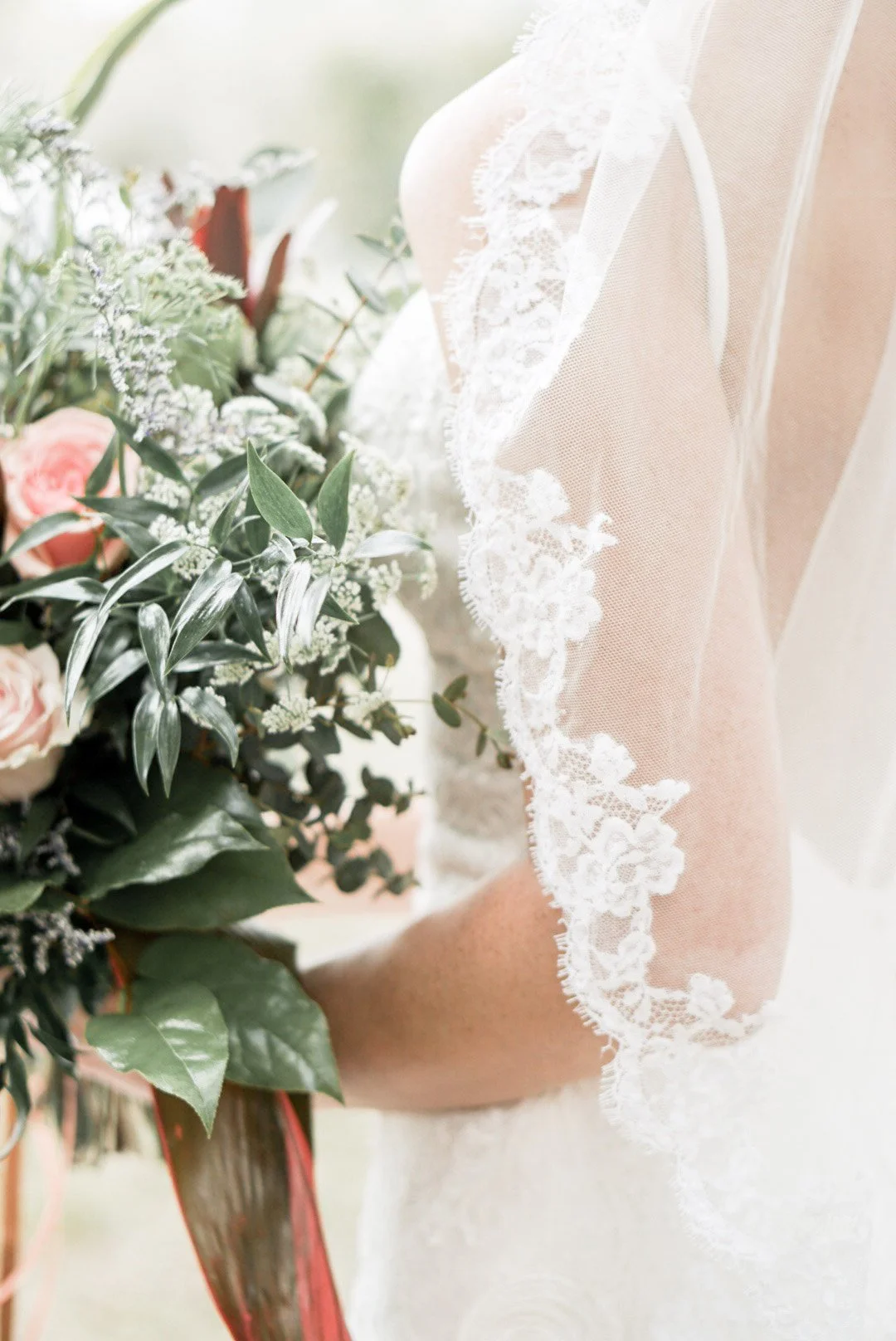 Close-up of a bride holding a bouquet of pink roses and greenery, wearing a white lace wedding dress with a decorative lace veil.