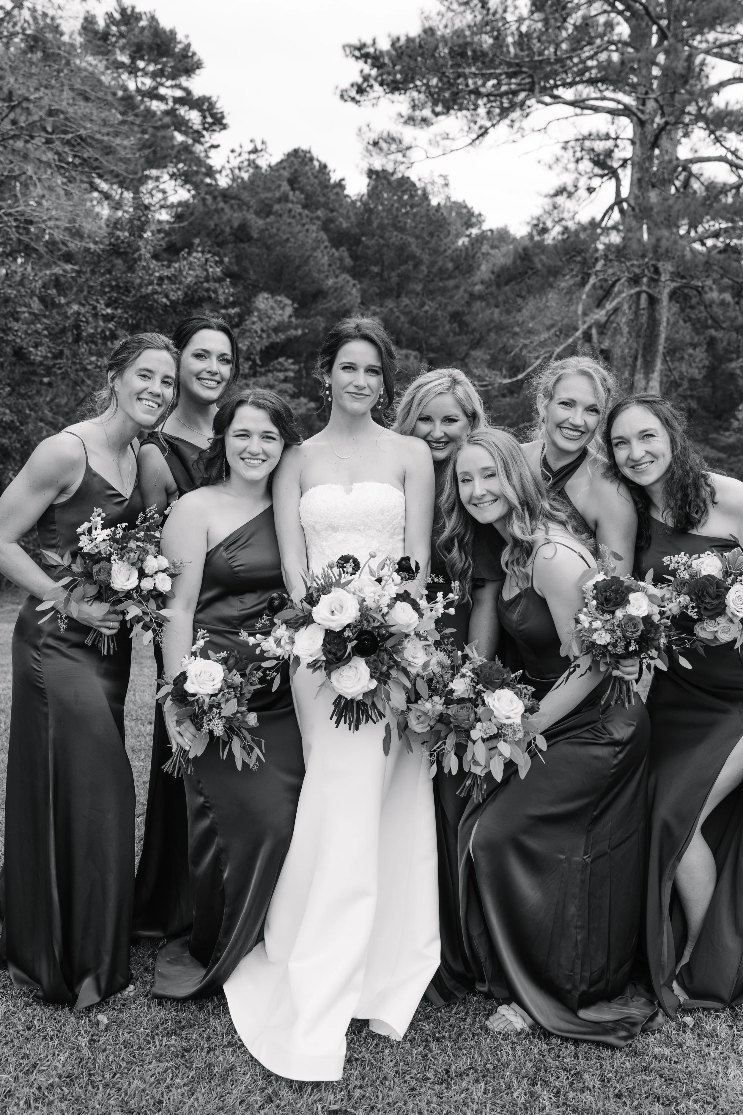 Black and white photo of a bride in a white wedding dress surrounded by seven bridesmaids in black dresses with floral bouquets outdoors in a natural setting with trees.