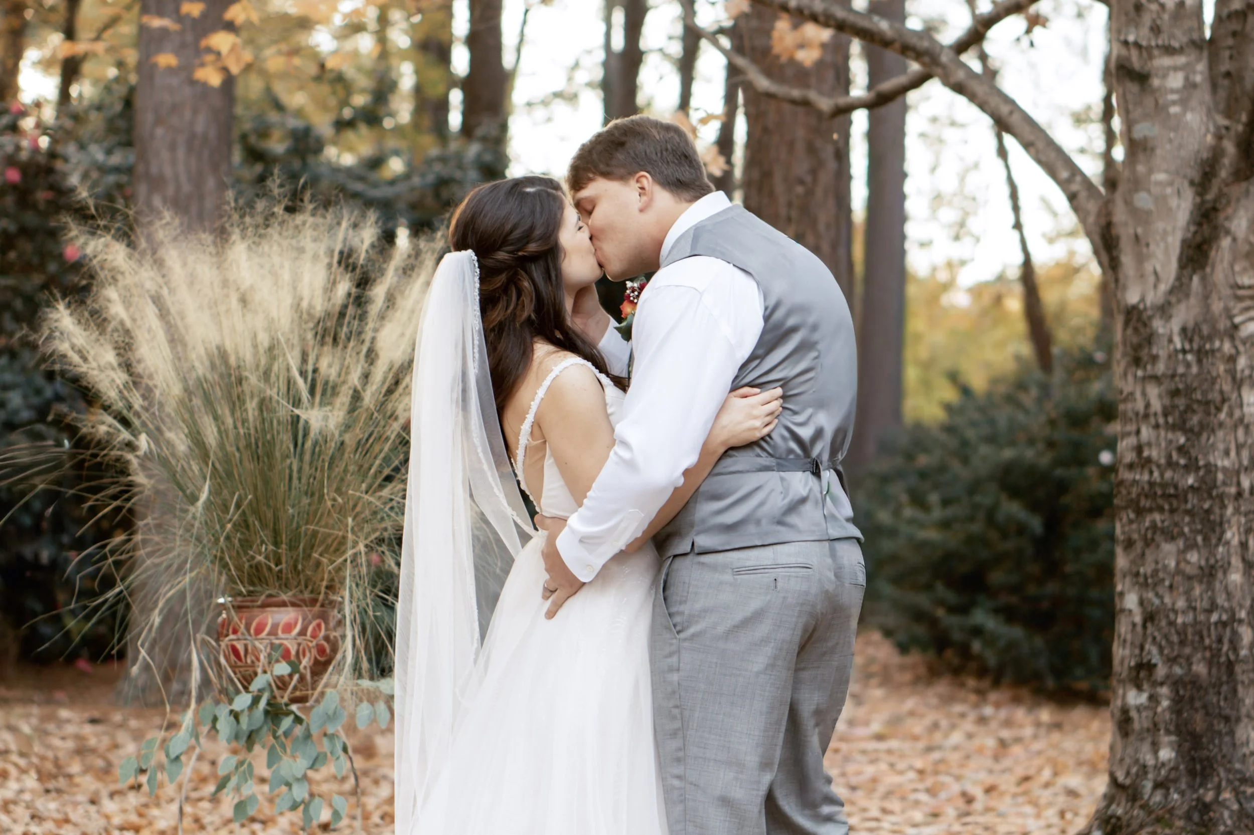 A bride and groom kissing outdoors among trees and fallen leaves at a wedding.
