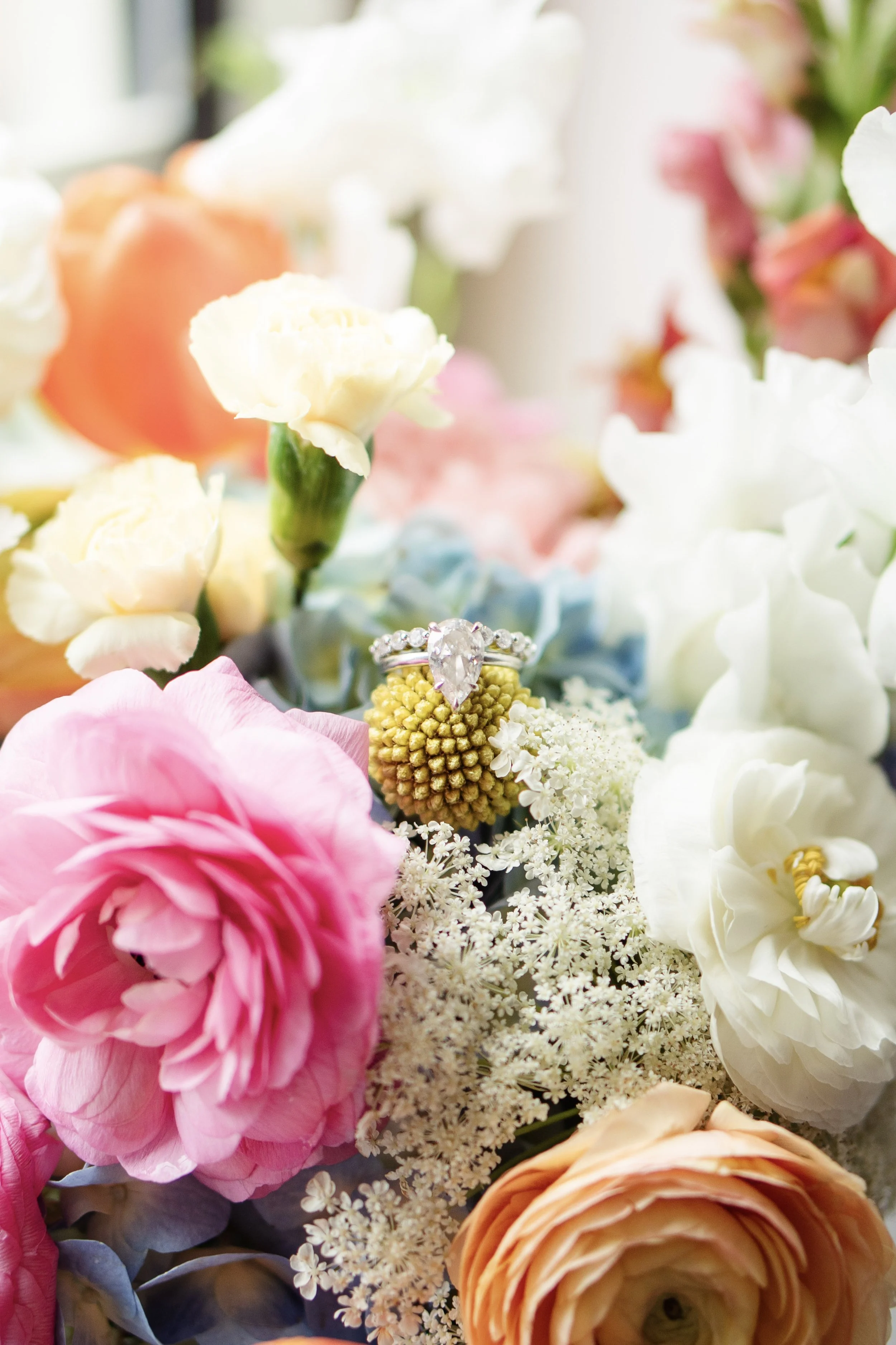 A close-up of a bouquet of colorful flowers with a diamond engagement ring placed on a small yellow flower in the center.