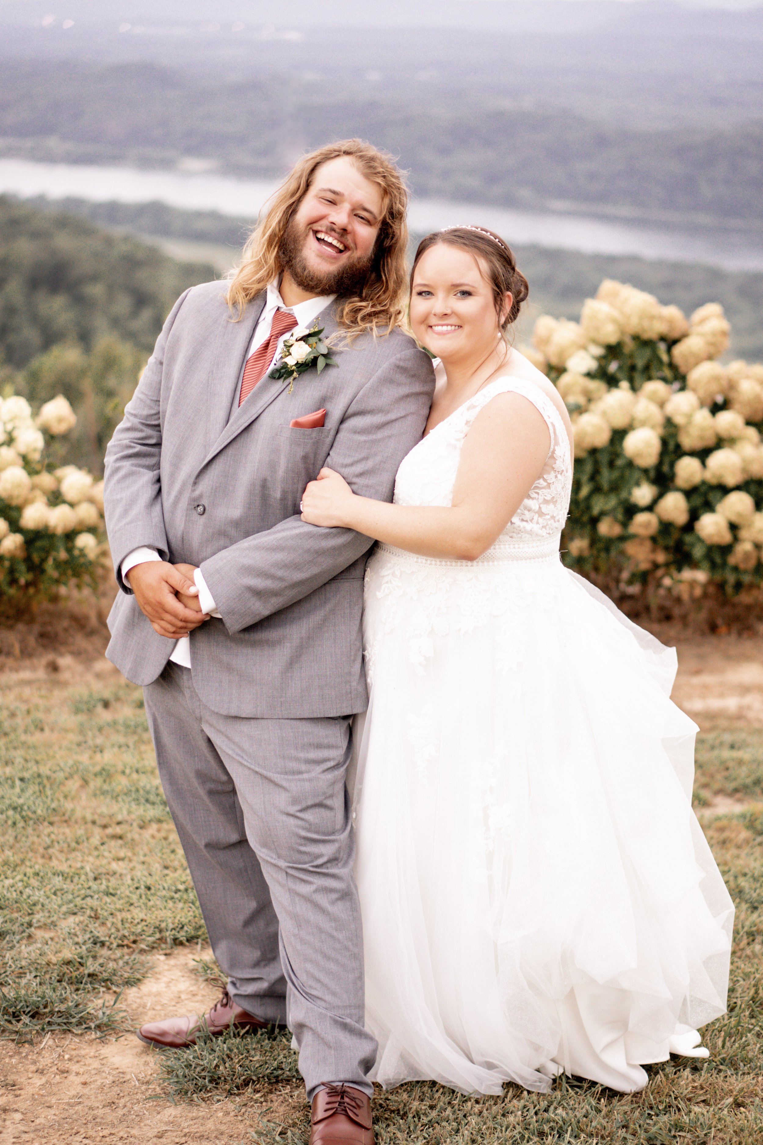 A happy newlywed couple standing outdoors, smiling, with a scenic landscape and blooming bushes in the background, dressed in wedding attire.