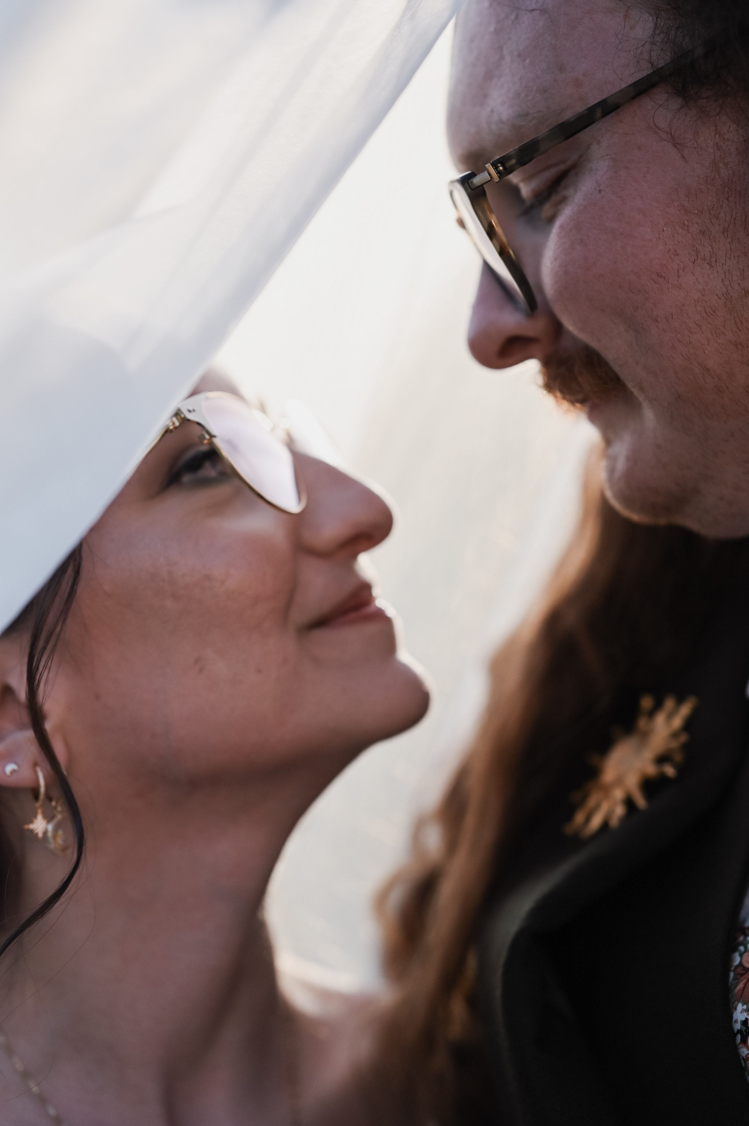 A close-up of a couple facing each other, both wearing sunglasses, with white and tan-colored hats. The woman has dark hair, earrings, and subtle makeup, and the man has a mustache, glasses, and long hair. They are outdoors, with a soft, natural back