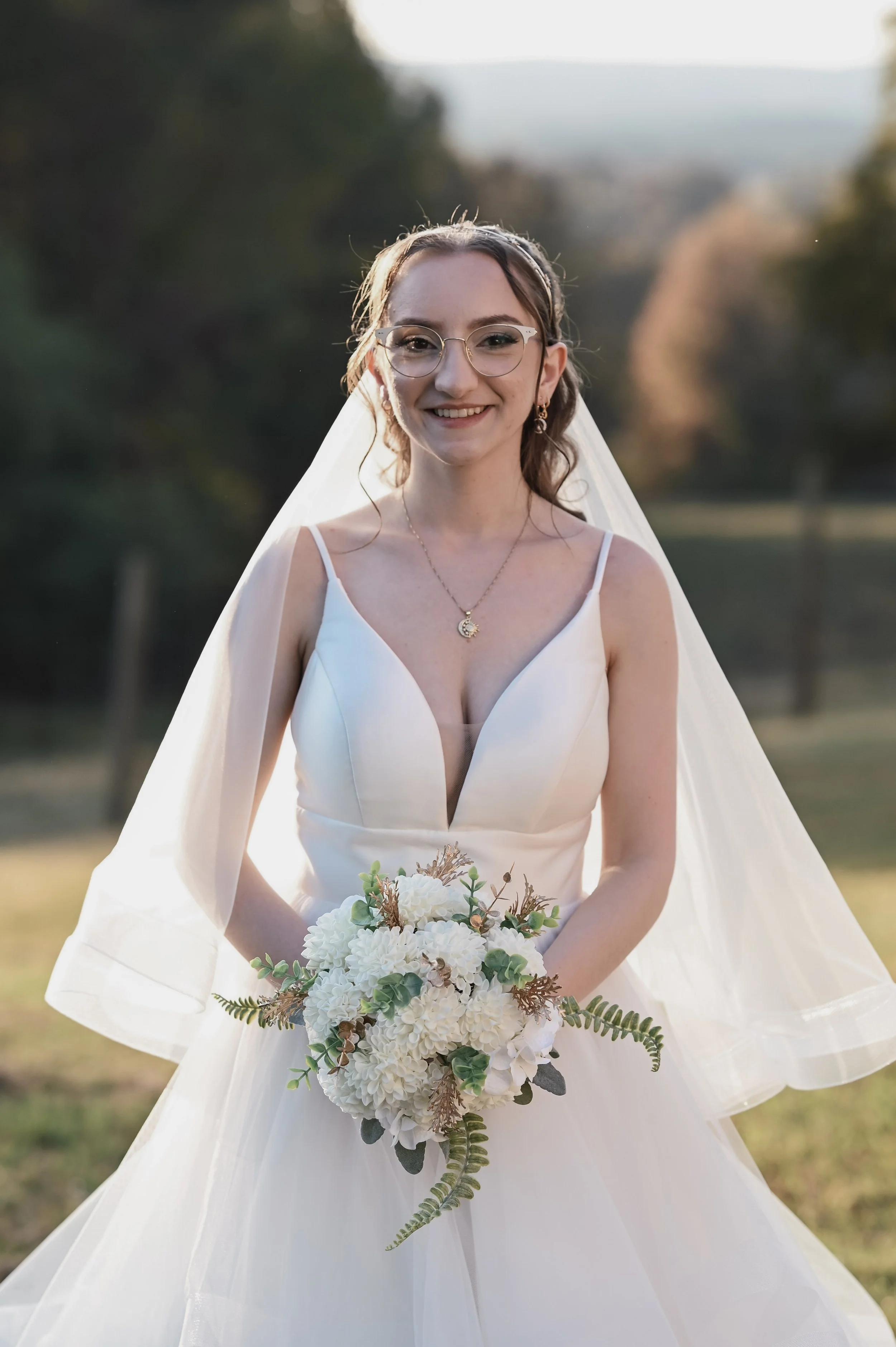 A smiling bride in a white dress holding a bouquet outdoors.