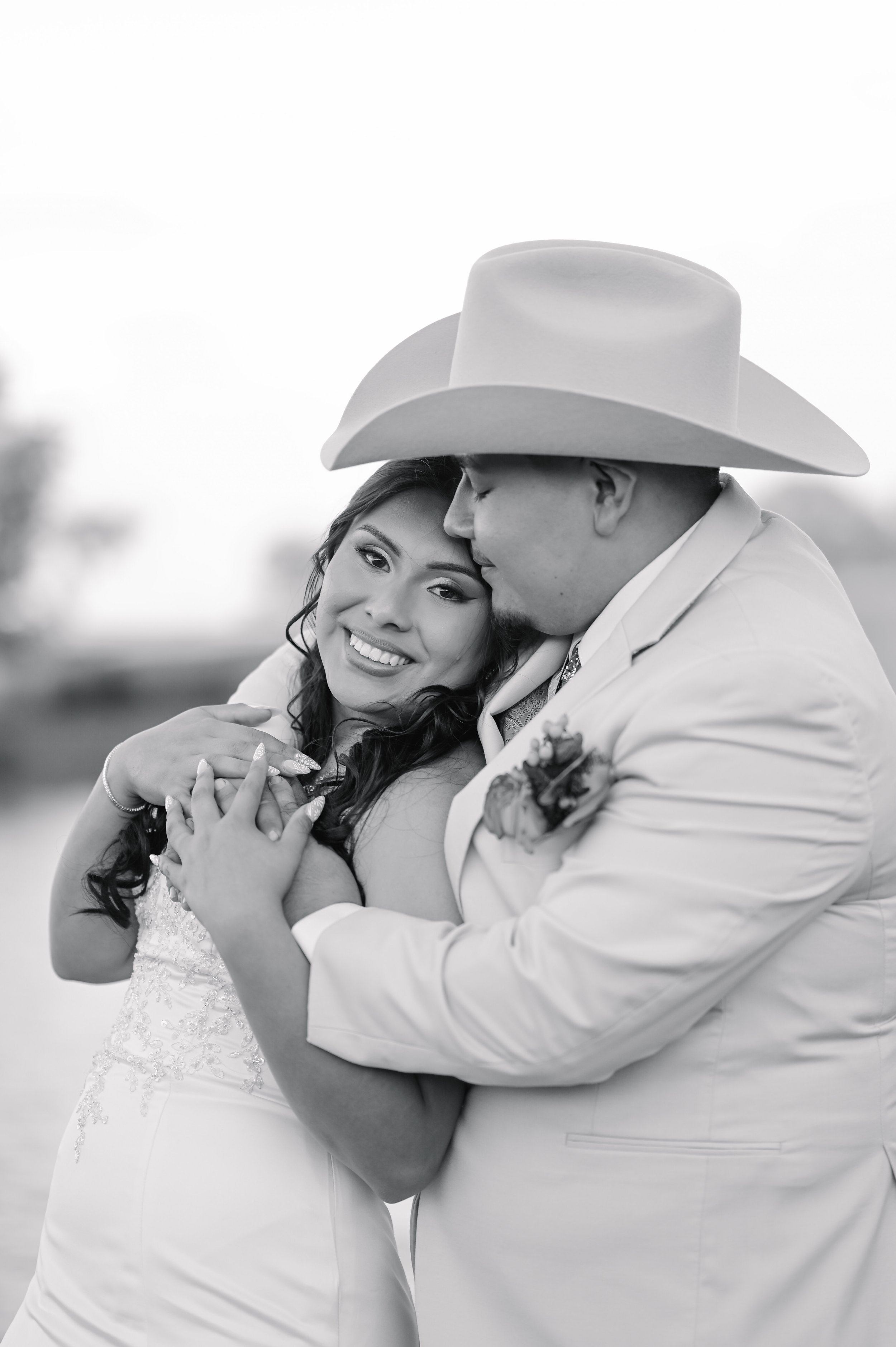 A black-and-white photo of a couple embracing each other, with the woman smiling and the man wearing a large cowboy hat kissing her on the forehead.