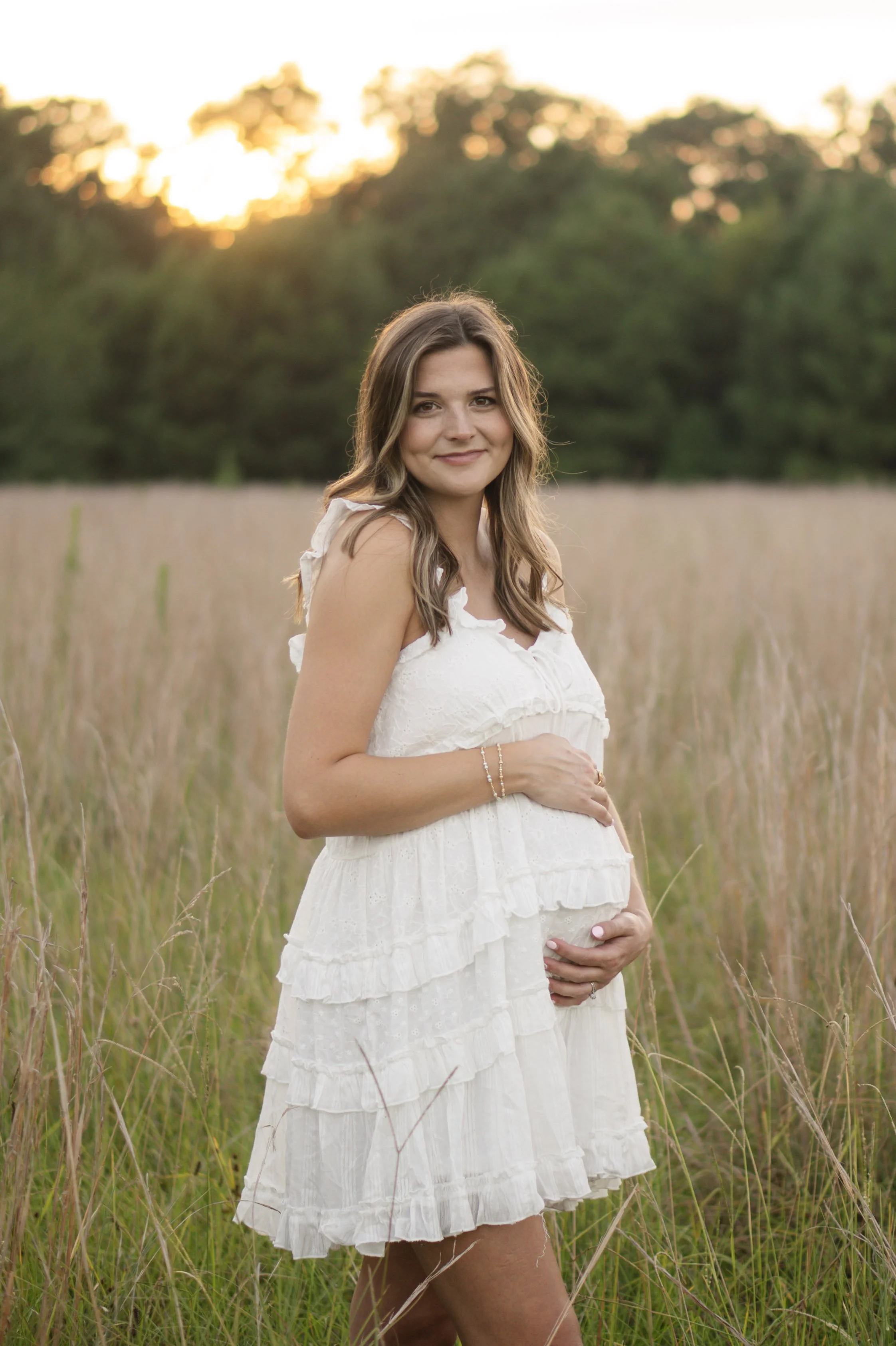 A pregnant woman standing in a grassy field during sunset, wearing a white dress and smiling, with her hand on her belly.