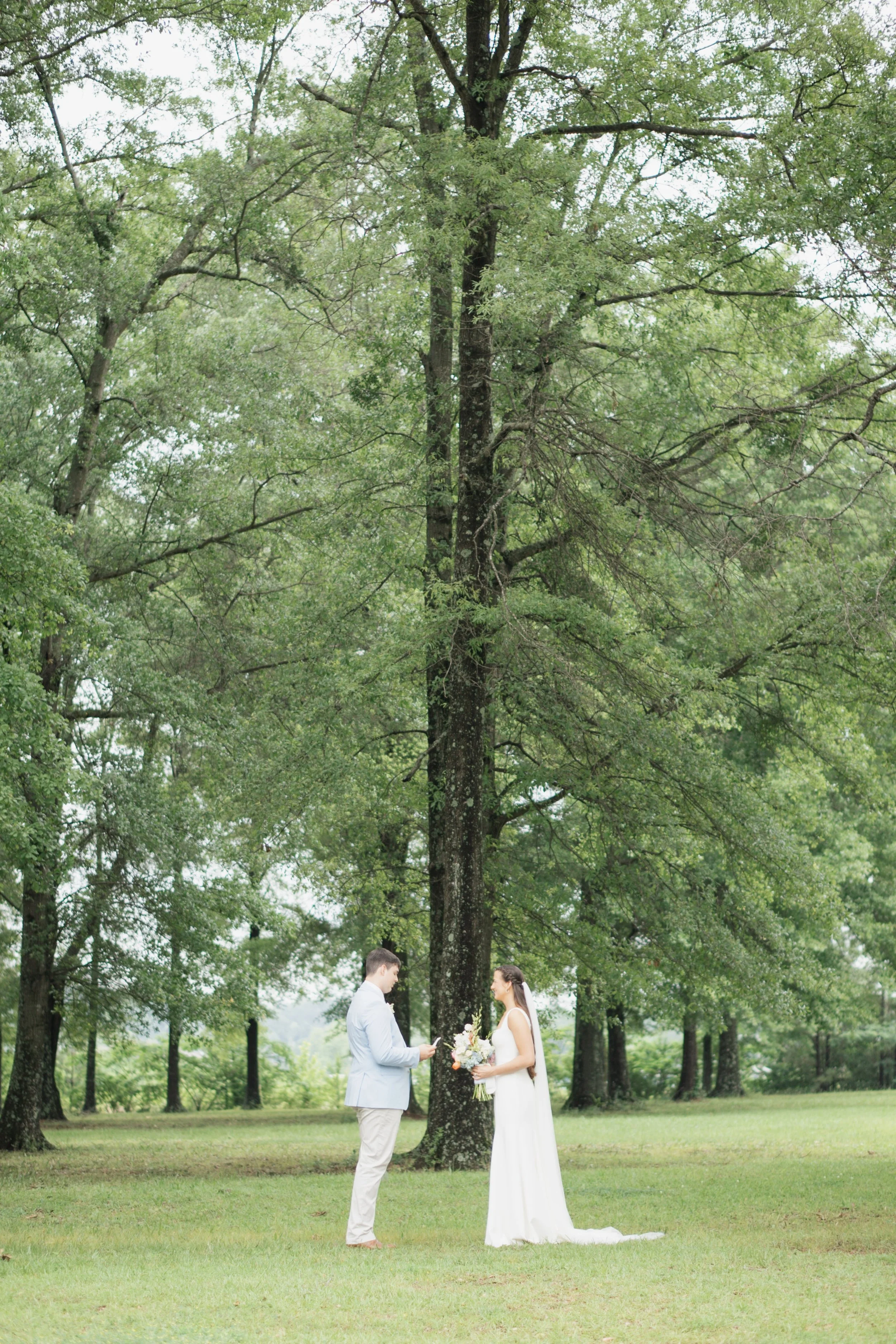 A wedding ceremony taking place outdoors under a large tree, with a bride in a white dress holding a bouquet and a groom in a light blue suit standing nearby.