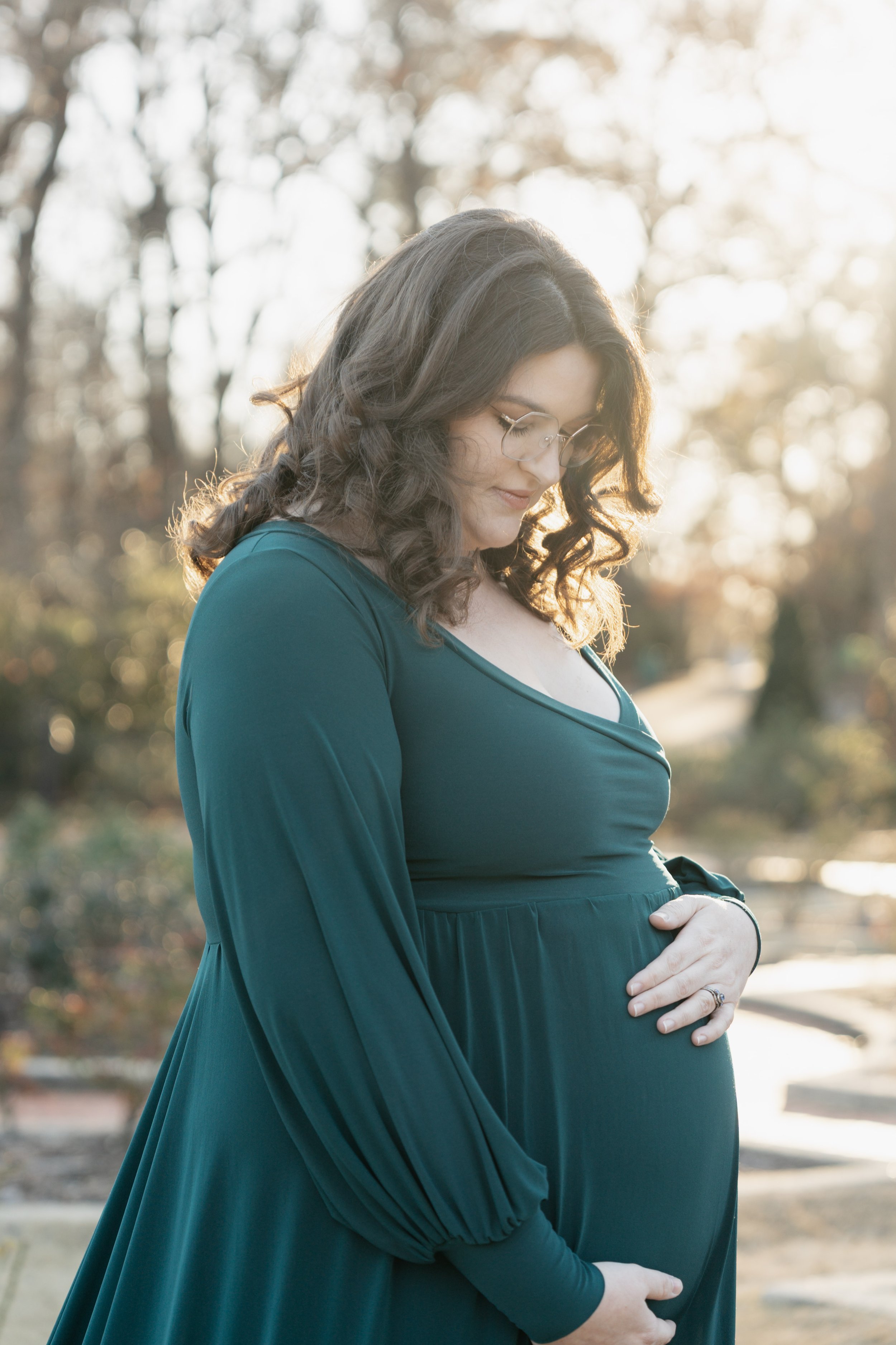 A pregnant woman with curly hair and glasses standing outdoors at sunset, gently touching her baby bump.